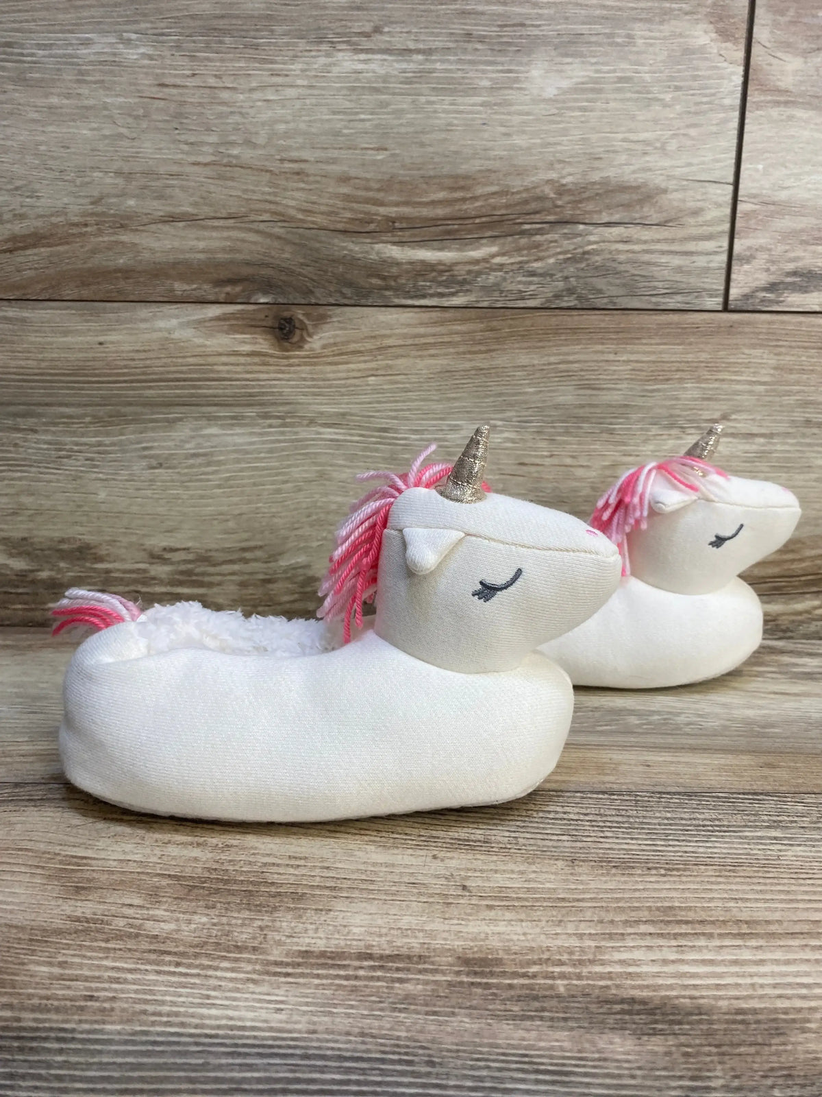 Side view of Gap Cozy Unicorn Slippers in white size 9/10c showcasing gold horn pink yarn mane and embroidered eyes on wood plank floor