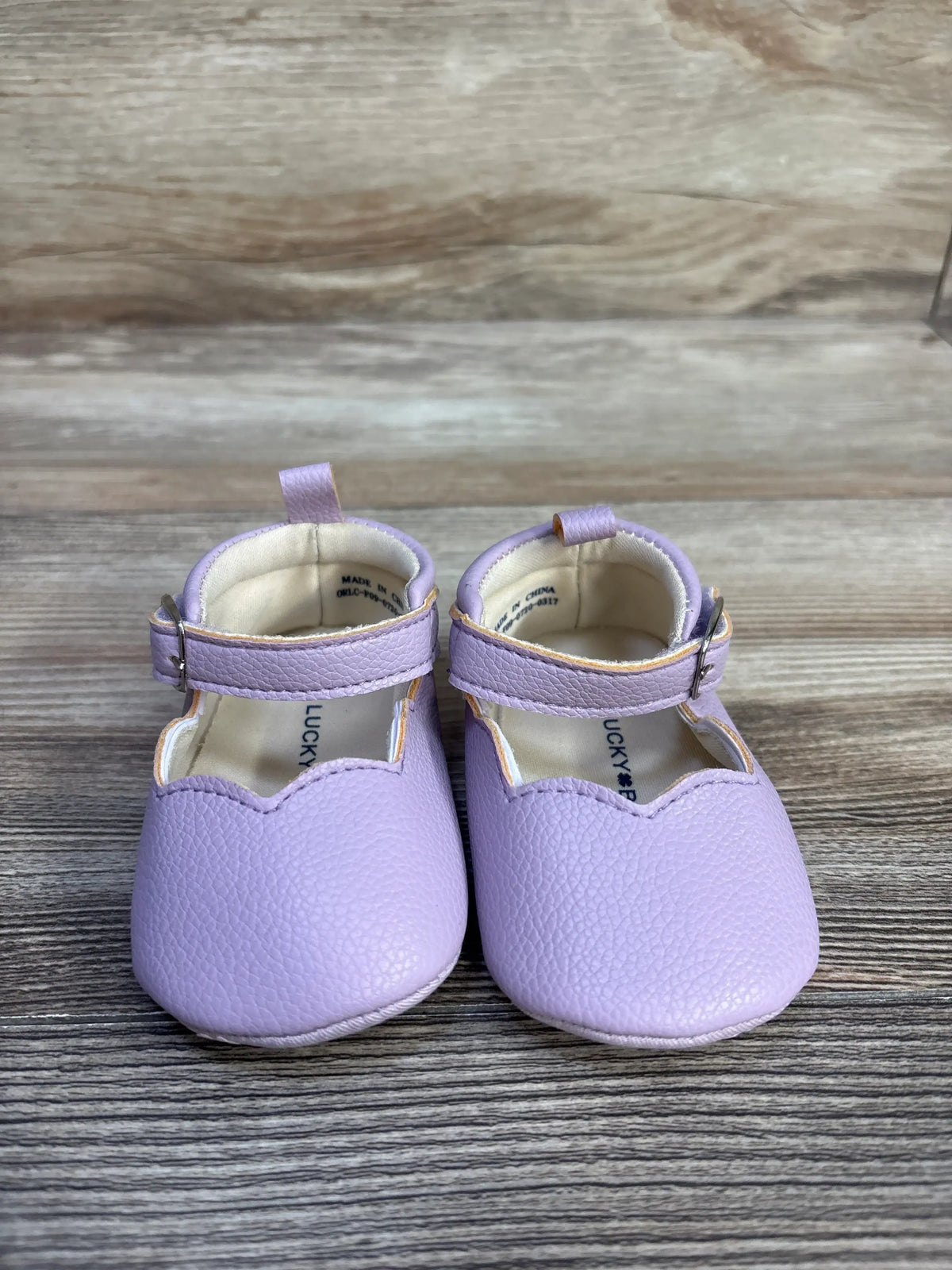 Front view of Lucky Brand purple Mary Jane crib shoes showcasing hook and loop buckle closures and pull tabs on wood grain