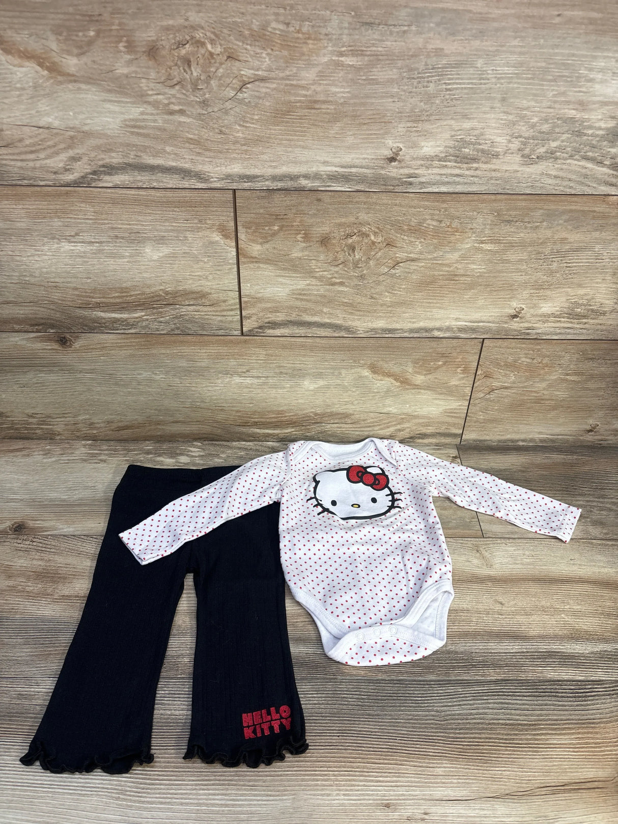 Flat lay of Hello Kitty 2-piece: white polka-dot bodysuit with Hello Kitty face and black ruffle-hem pants, 12 months
