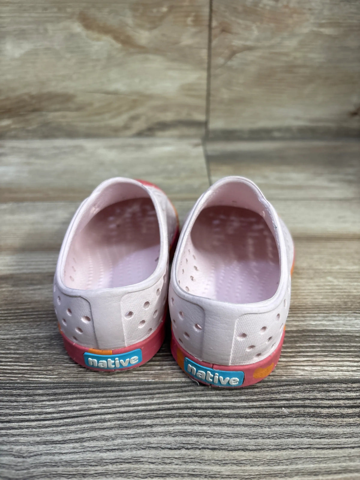 Rear view of pink Native Jefferson slip-on shoes, size 8C, showing perforated uppers and Native heel branding on wood floor
