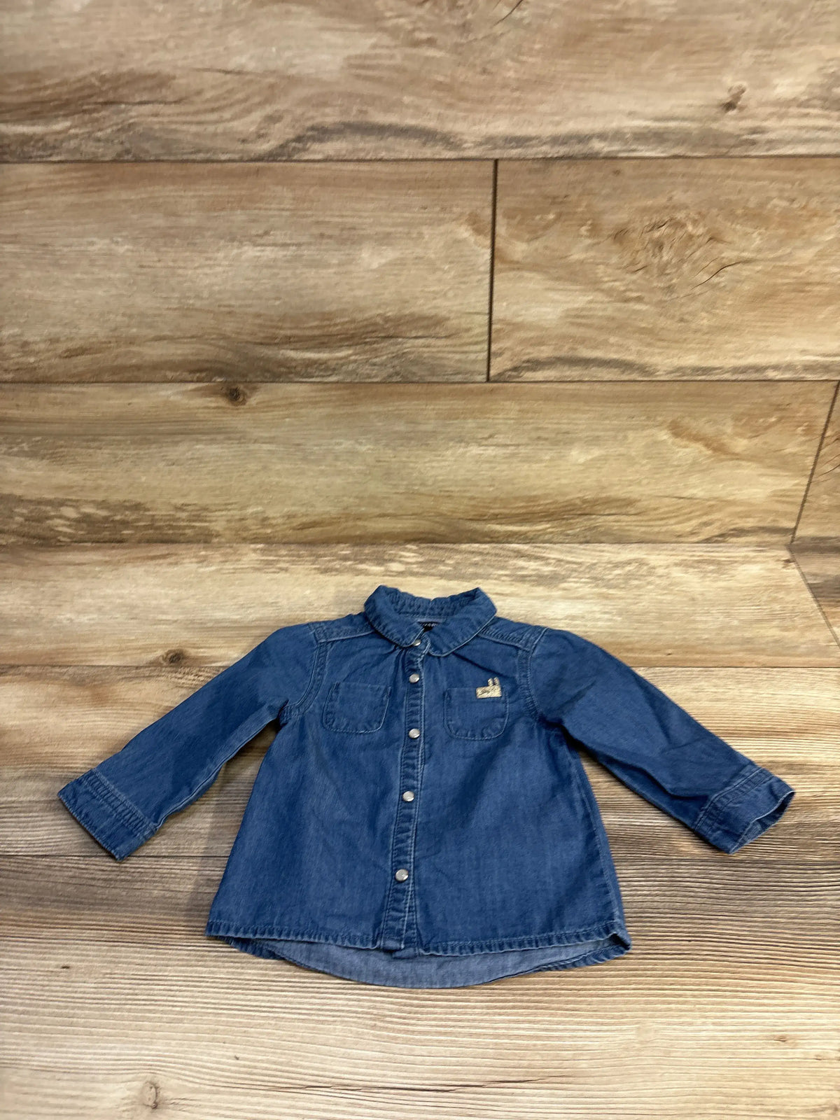 Front view of chambray long-sleeve shirt, size 12M, showing chest pockets and snap buttons on wood background