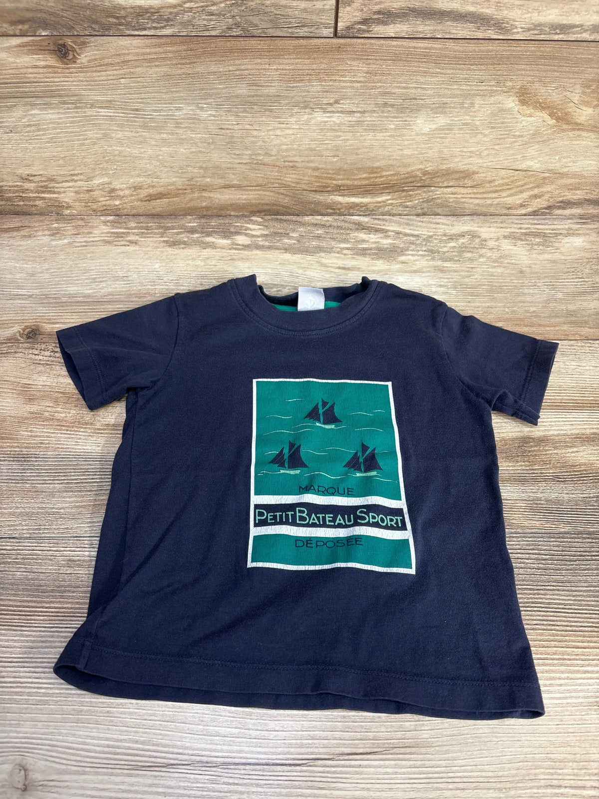 Front view of a navy Petit Bateau short sleeve shirt showcasing a sailing graphic and Petit Bateau Sport text with a wood background