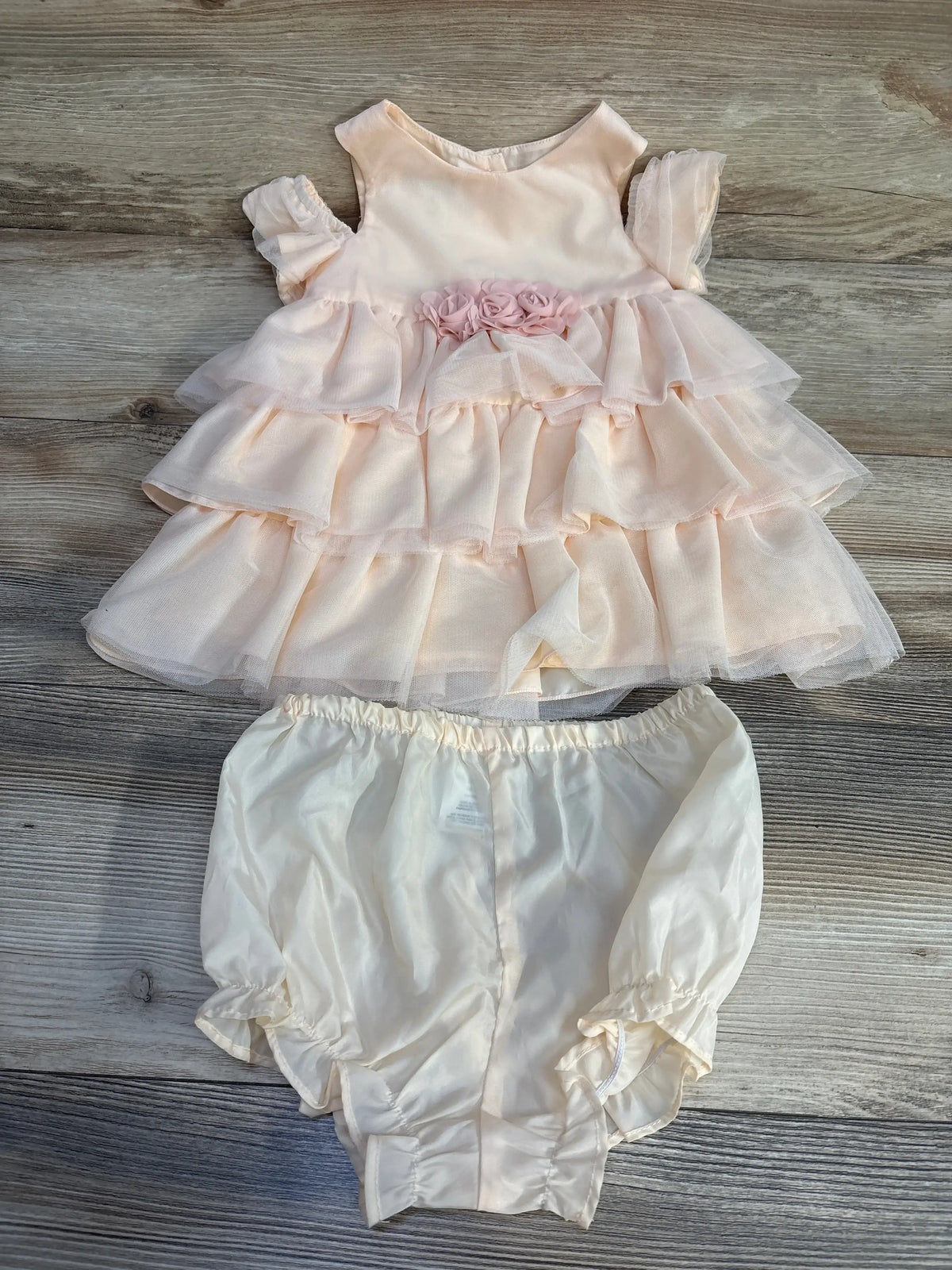 Top down view of peach tiered baby dress showing ruffled tiers and rosette trim with cream bloomers on wood surface
