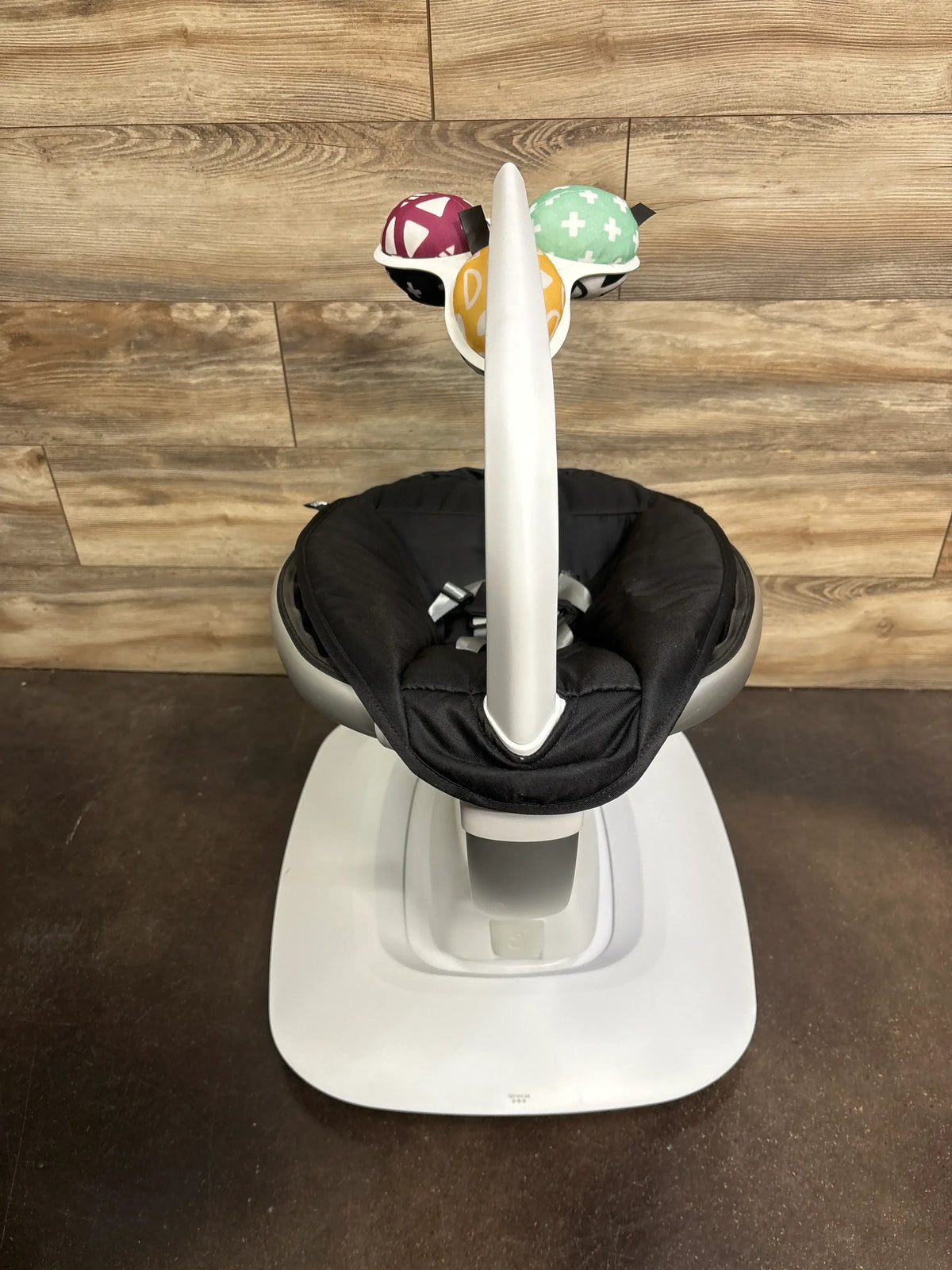 Front view of 4moms mamaRoo baby swing showcasing black padded seat and toy mobile with wood-paneled background