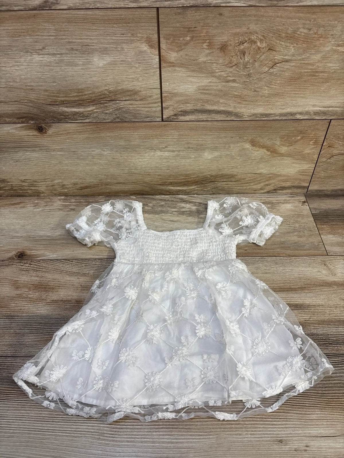 Flat lay of white embroidered tulle toddler dress showing smocked bodice, puff sleeves and floral tulle overlay on wood floor