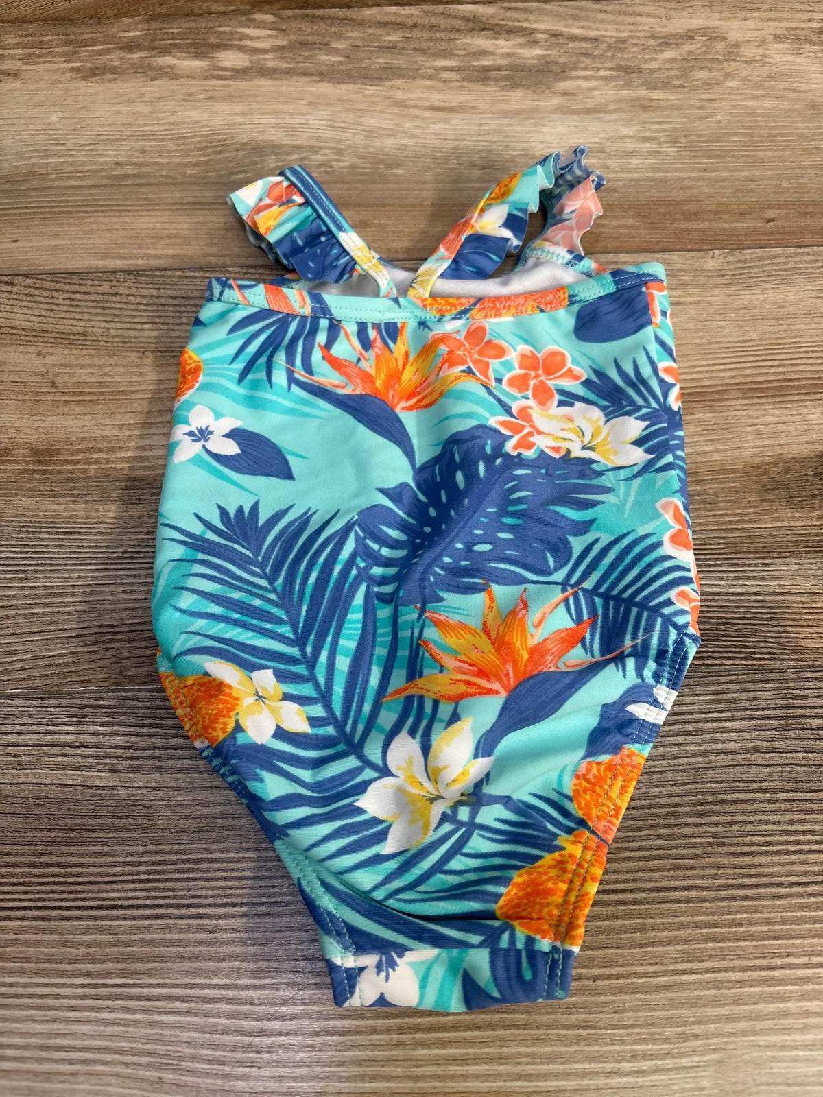 Top-down view of newborn 0–3M one-piece swimsuit with turquoise tropical floral and pineapple print and ruffle shoulder straps on wood