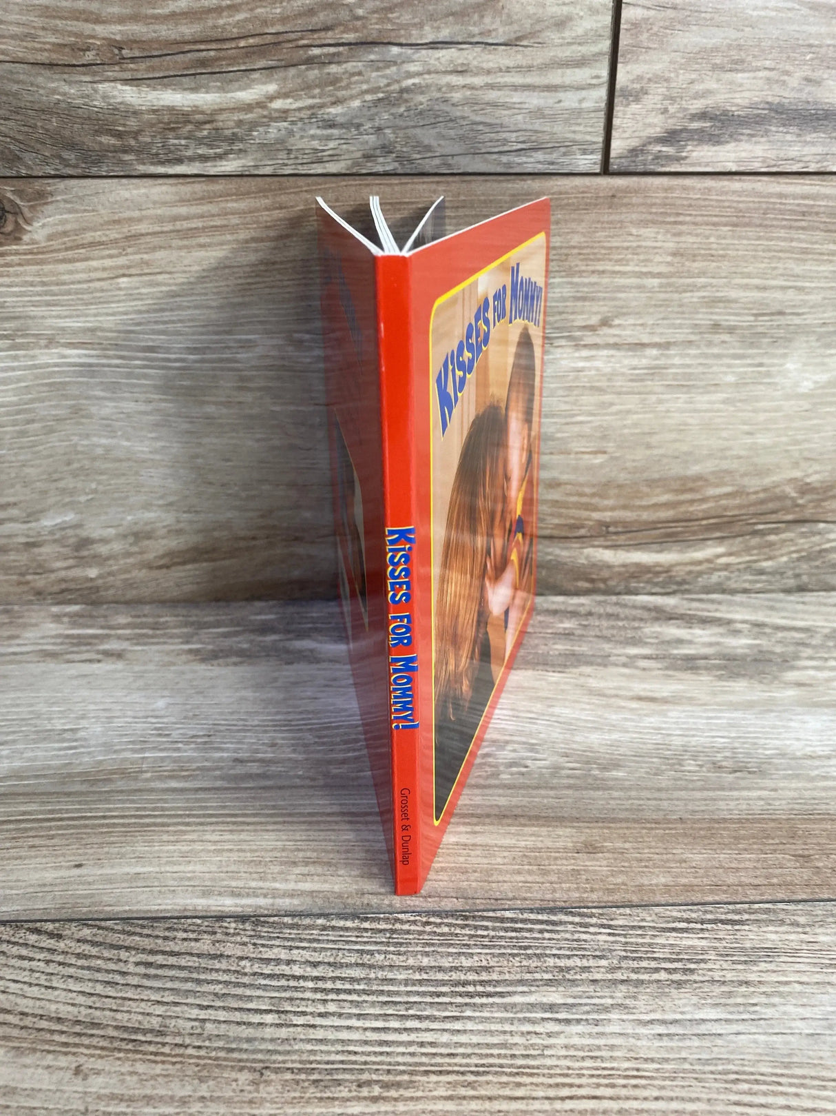 Three-quarter view of Kisses for Mommy board book showcasing red cover spine and front edge propped on wood plank floor