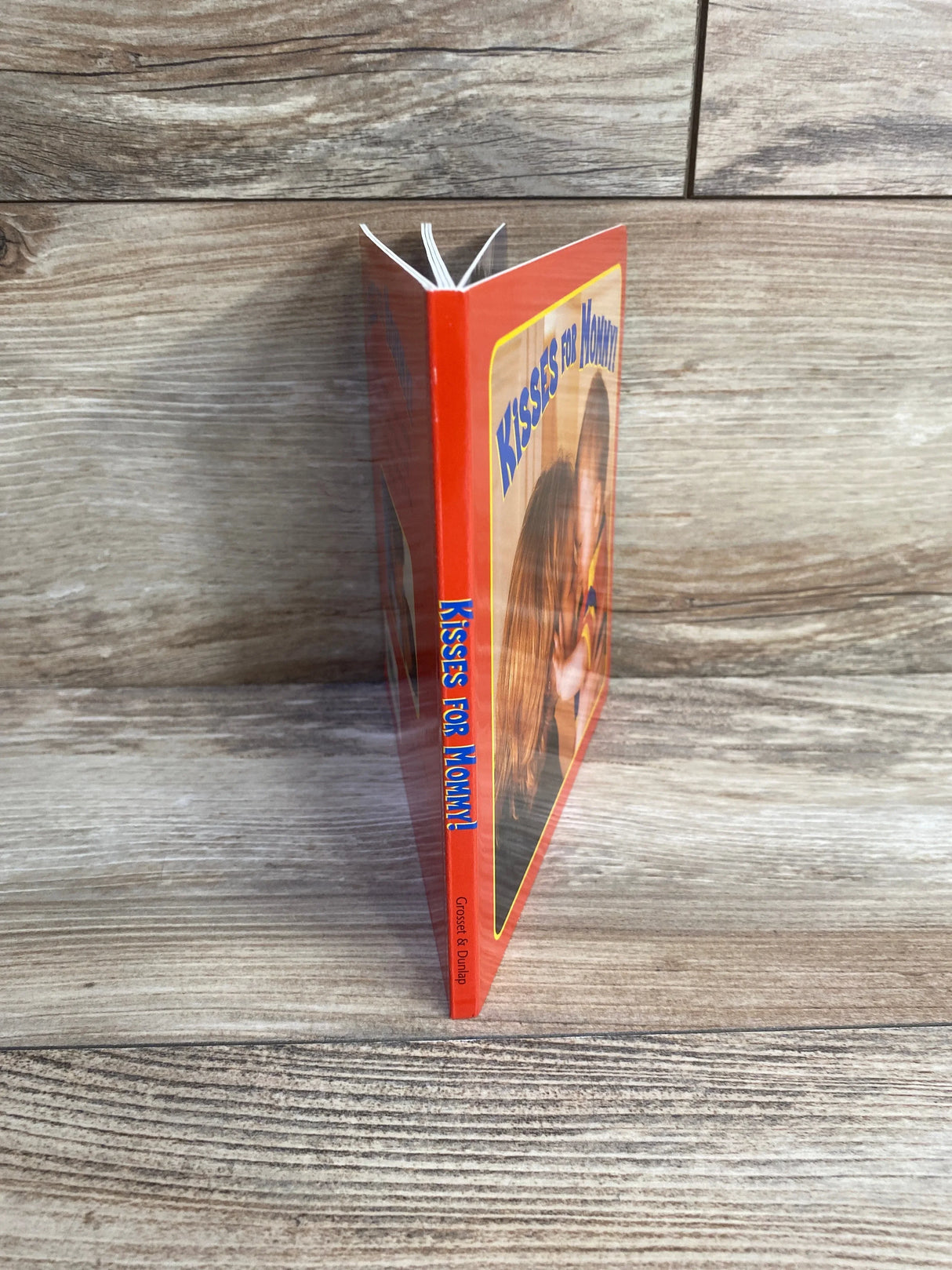 Side view of Kisses for Mommy! board book with orange spine and cover showing mother kissing child on wood background