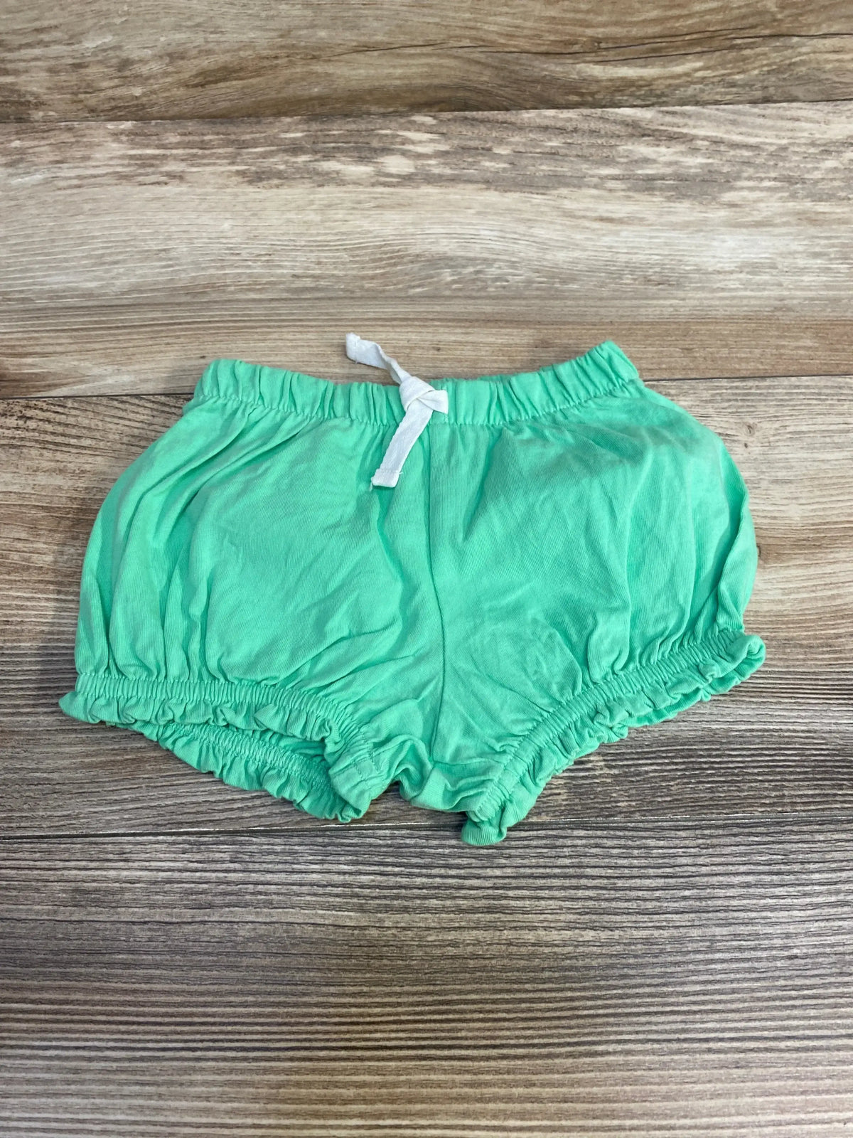 Flat lay of green baby shorts size 3 to 6 months showcasing elastic waistband with white drawstring and ruffled leg openings on wood surface