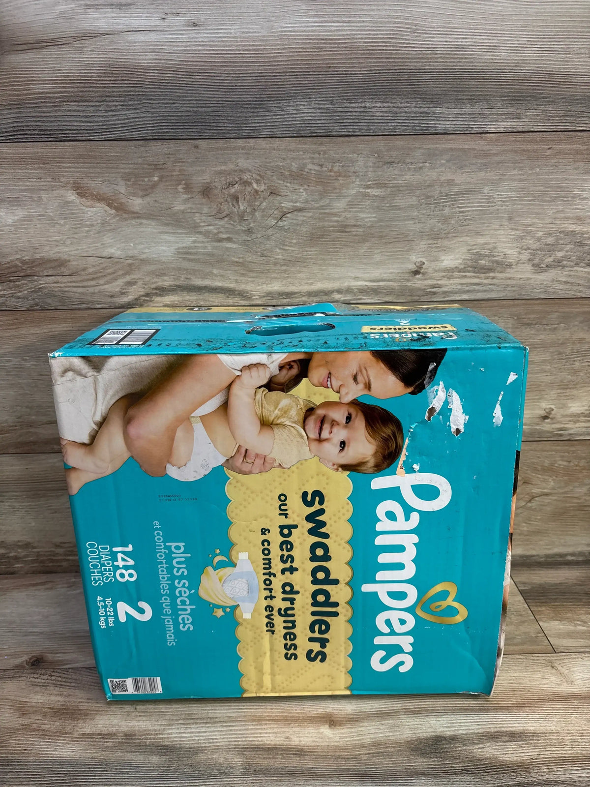 Angled top view of Pampers Swaddlers size 2 148-count diaper box showing packaging artwork with the text our best dryness visible and scuffed teal box on wood floor