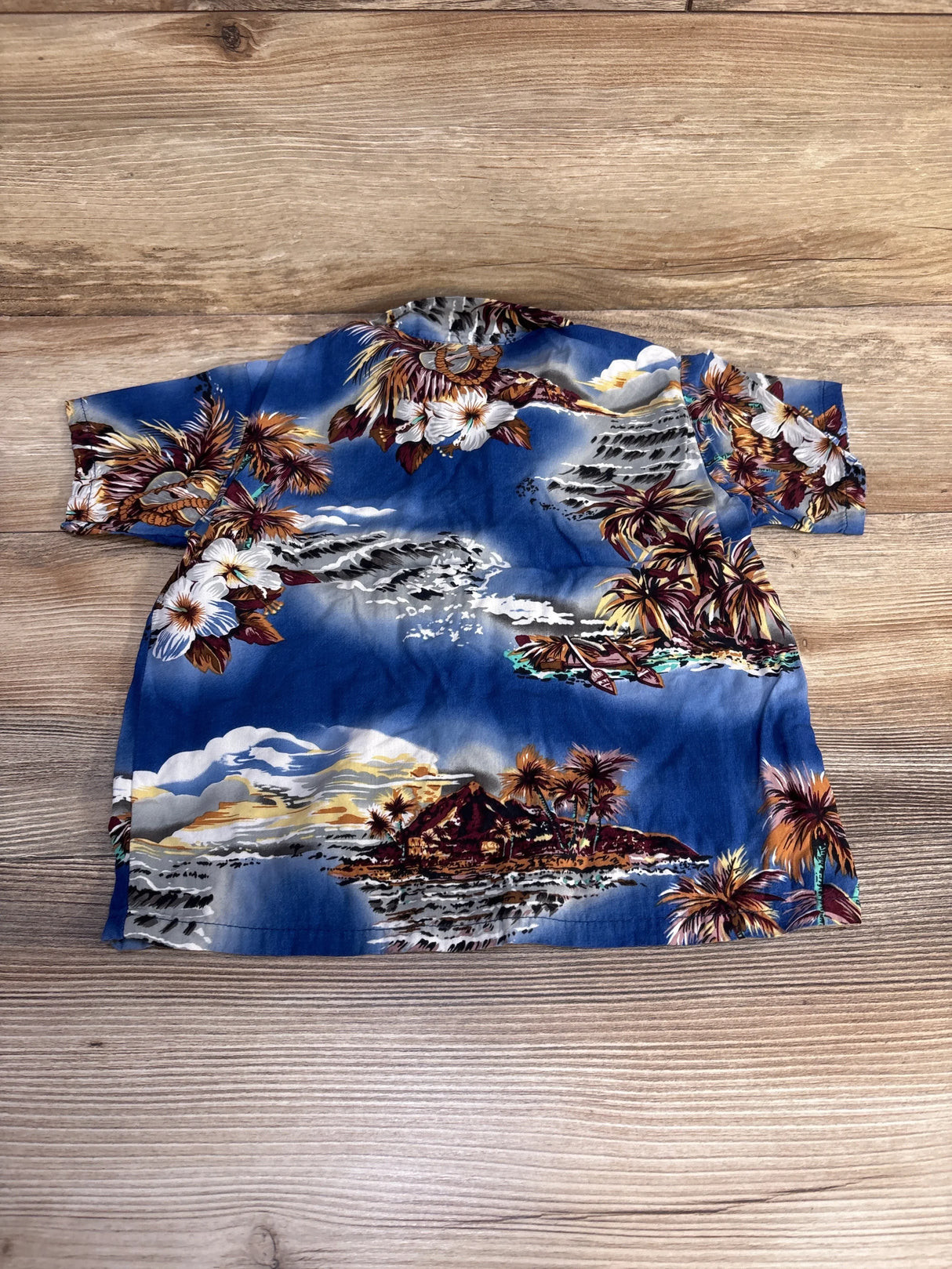Back view of blue Hilo Hattie 4T aloha button-up shirt showing tropical island and hibiscus print on light wood background