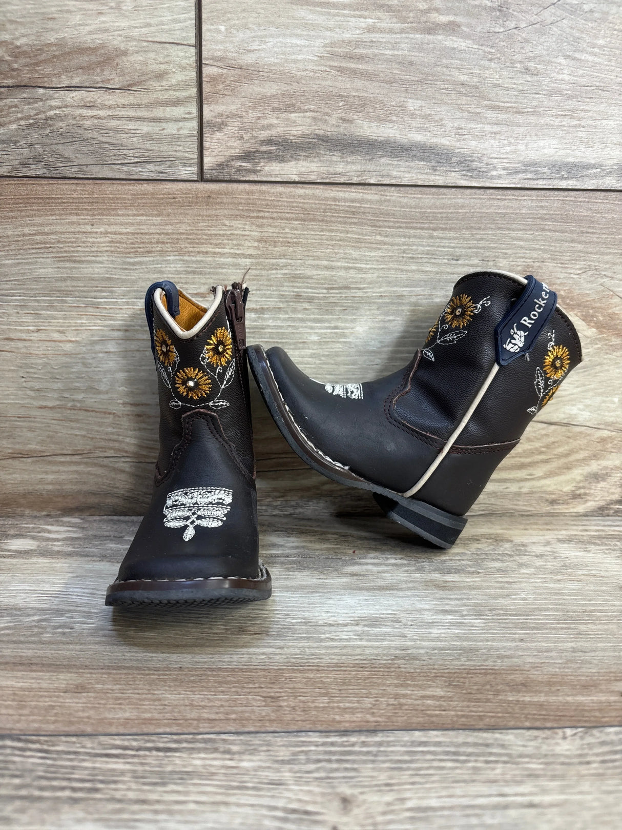 Three-quarter view of Rock’em toddler brown western boots with yellow sunflower embroidery and side zipper on wood background