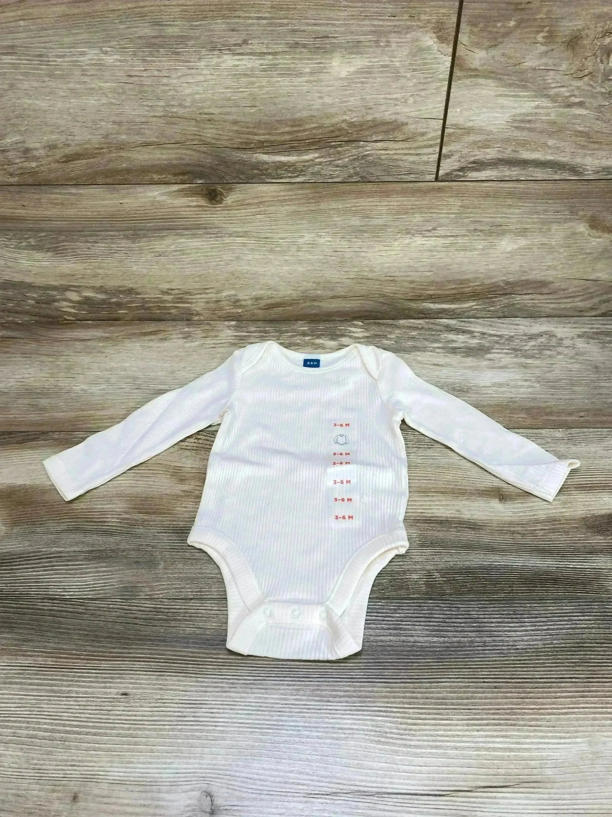 Flat lay of Old Navy ivory rib-knit long-sleeve baby bodysuit with snap crotch and printed 3-6M size tag on wood background