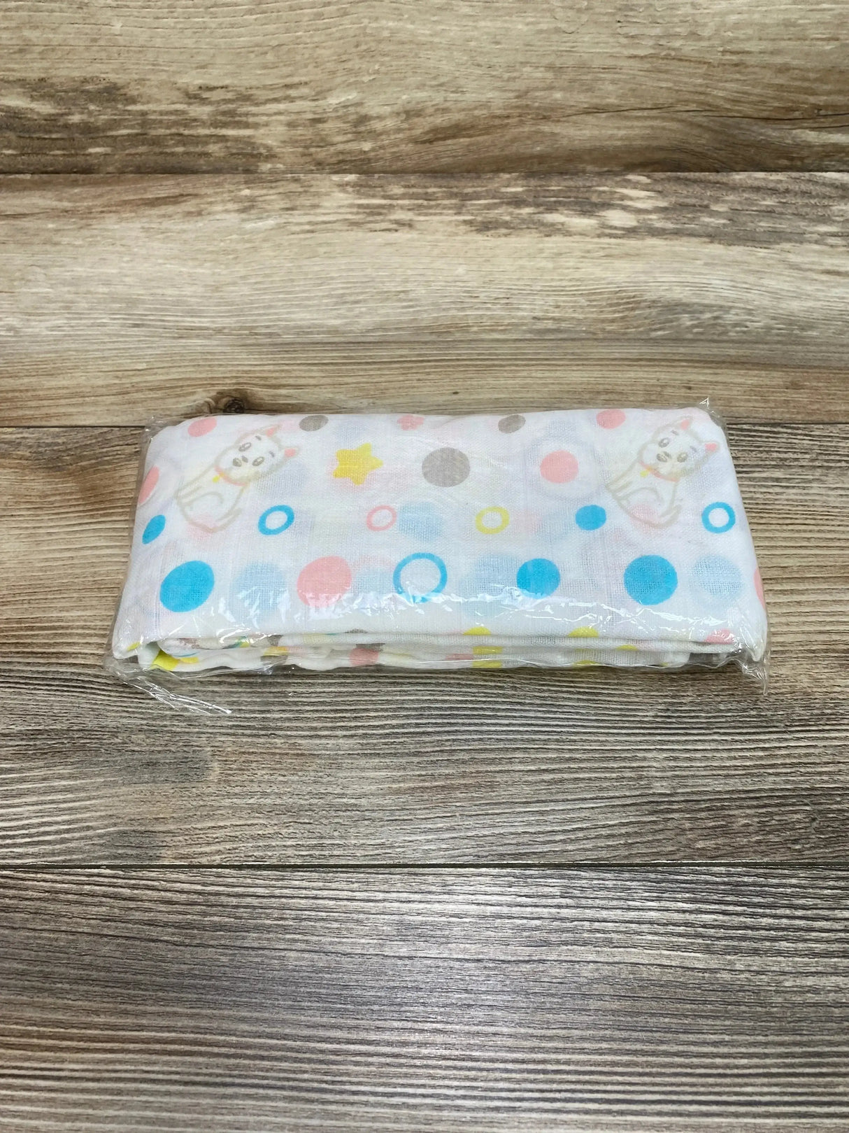 Folded Cucumberries cotton muslin swaddle blanket showing pastel paw and polka dot print in clear plastic packaging on wood background