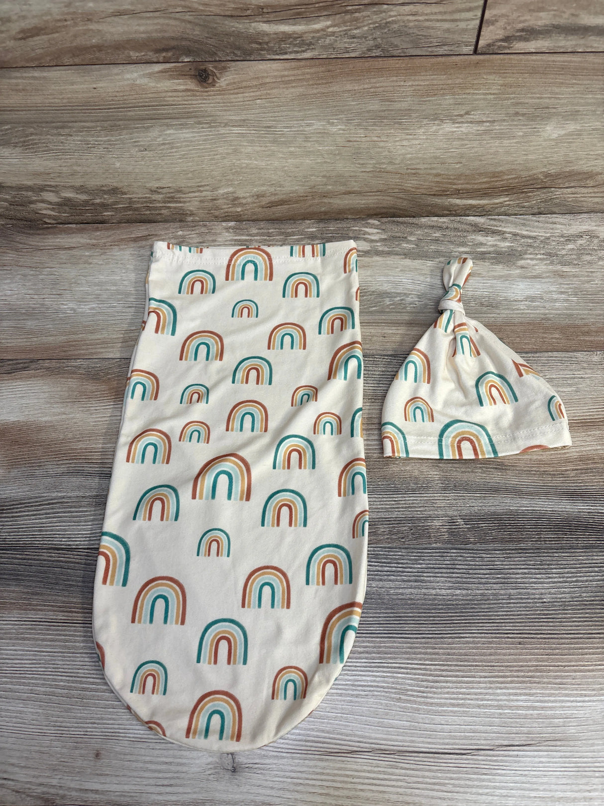 Cream baby swaddle and knotted hat set with teal and rust rainbow patterns on wood background