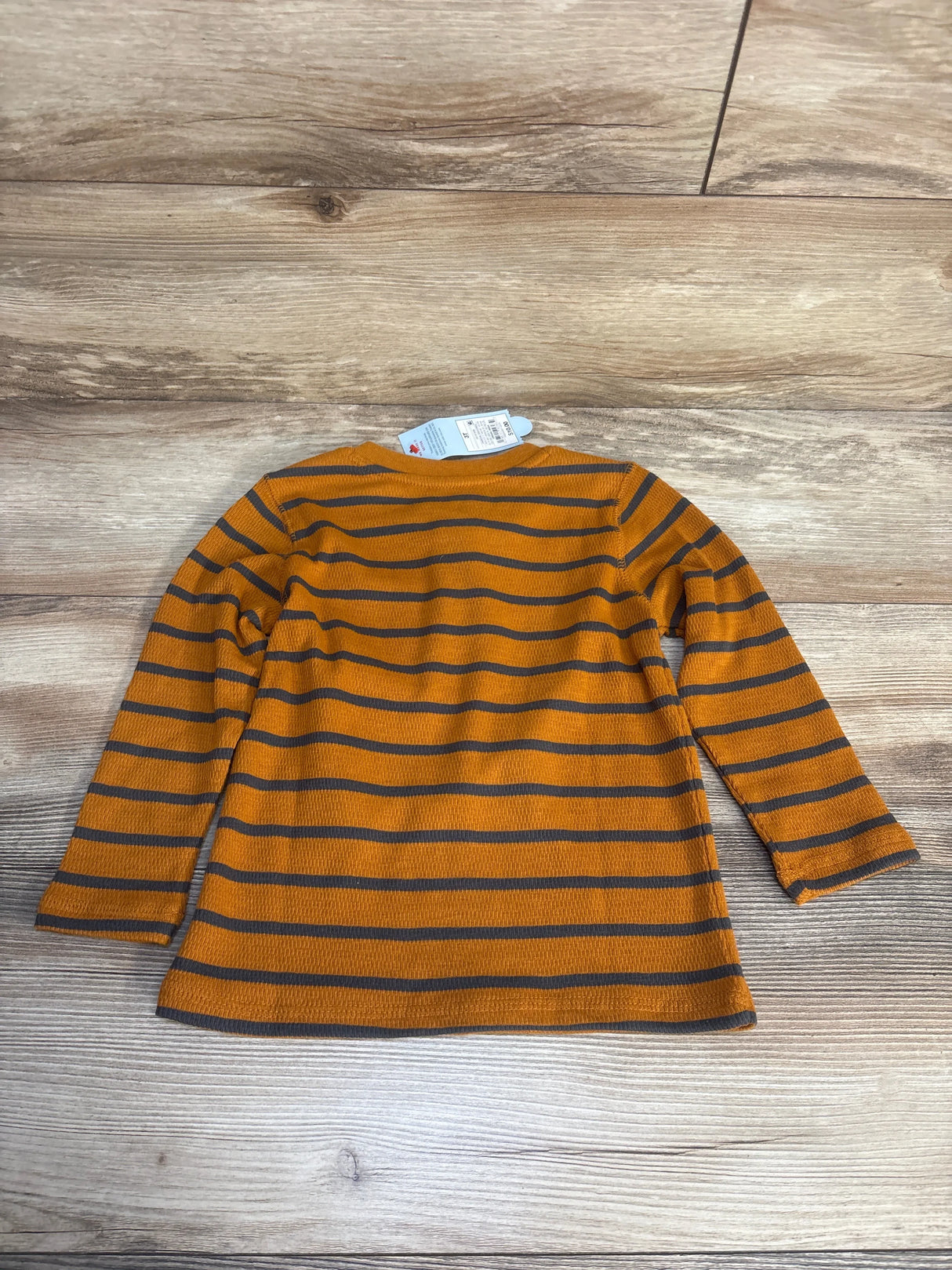 Kids' long sleeve shirt, orange with gray horizontal stripes, laid flat on wood floor