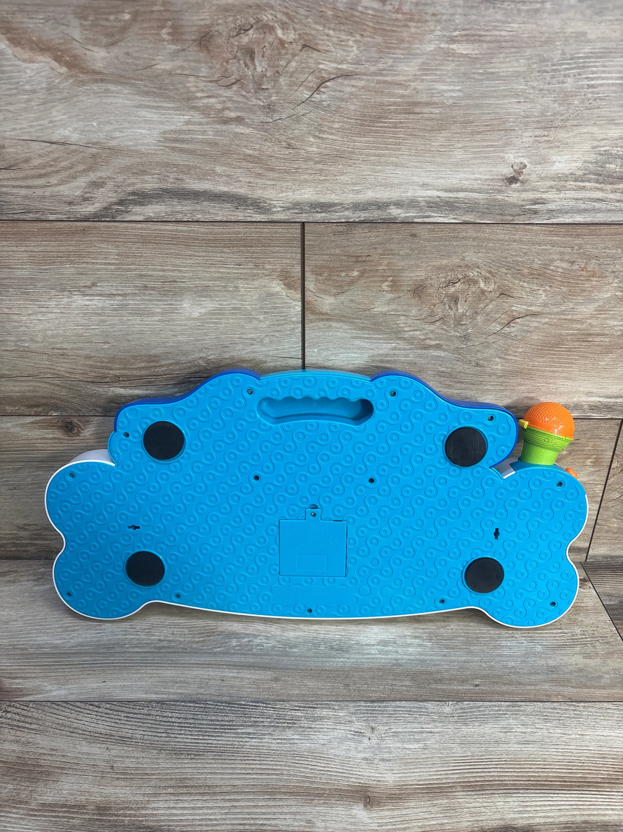 Bottom of Rolo's Keyboard toy showing blue textured base with battery door, black feet, and orange microphone on wood