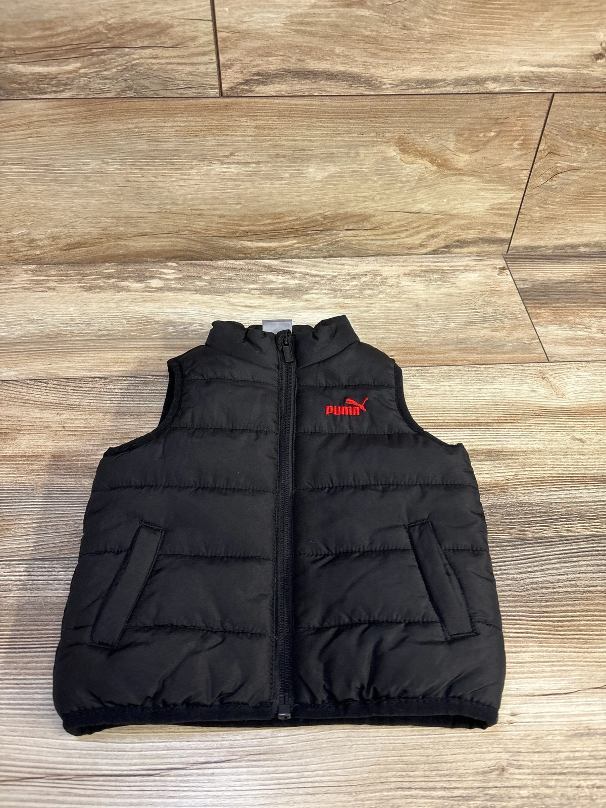 Front view of black Puma puffer vest size 12 months showcasing center zipper, red Puma logo, and two front pockets on light wood floor