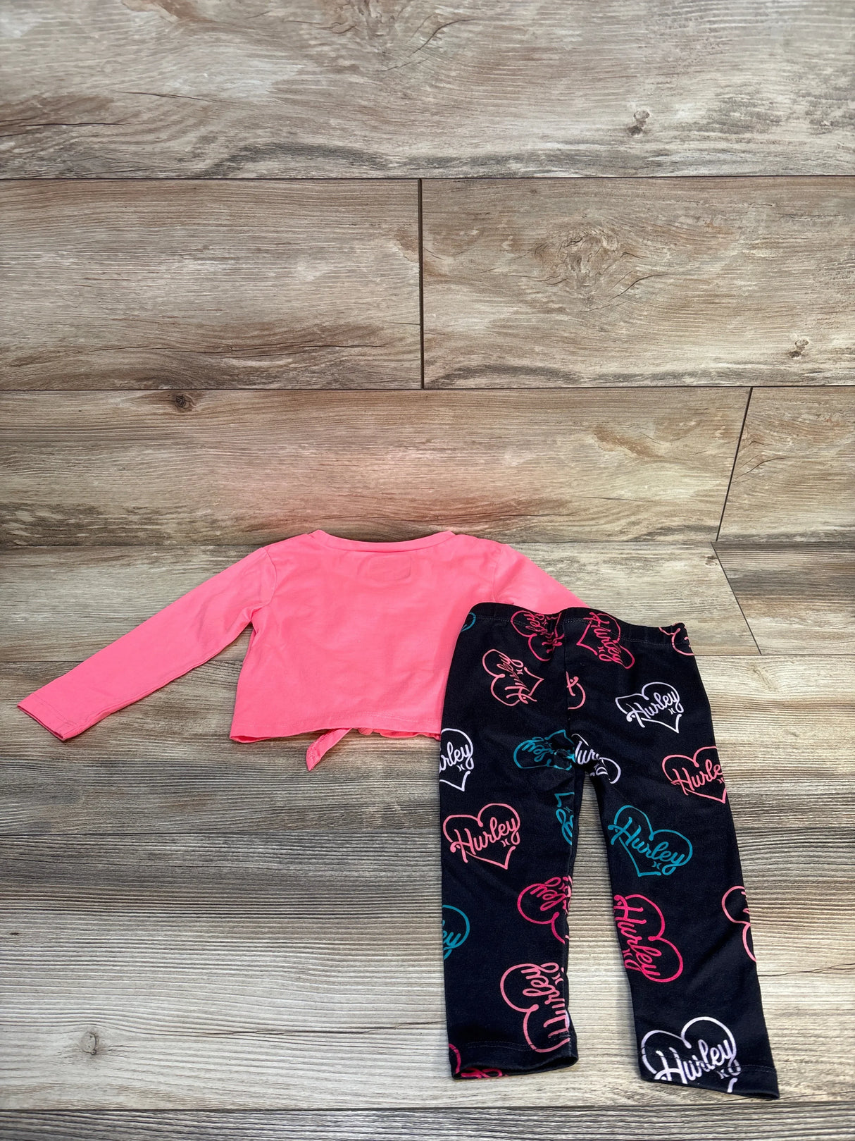 Flat lay of toddler Hurley 2pc set showcasing pink long sleeve top and black leggings with Hurley print on wood