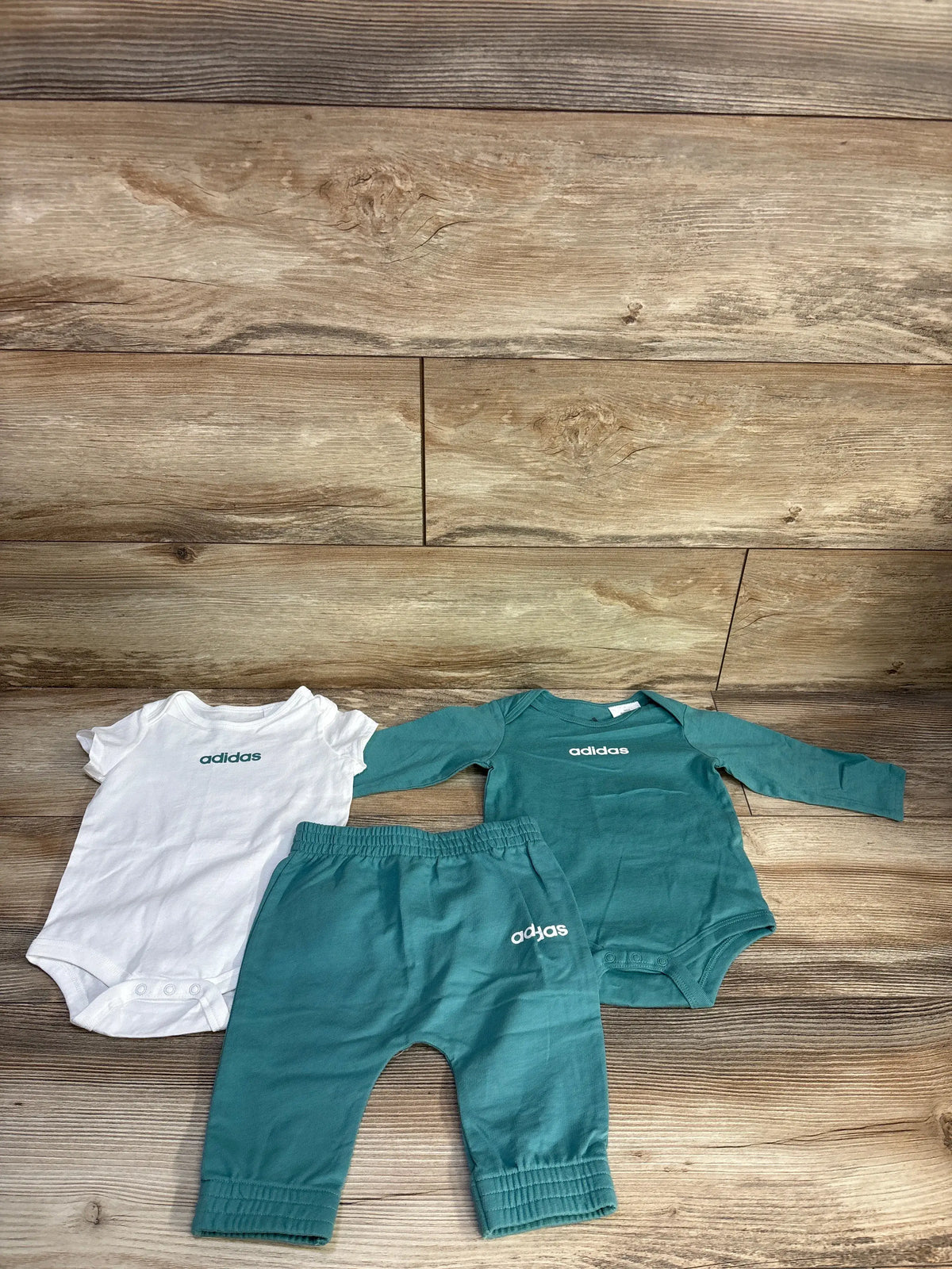 Front view of Adidas 3-piece bodysuit set showcasing green long-sleeve and white short-sleeve bodysuits with joggers on a wood surface