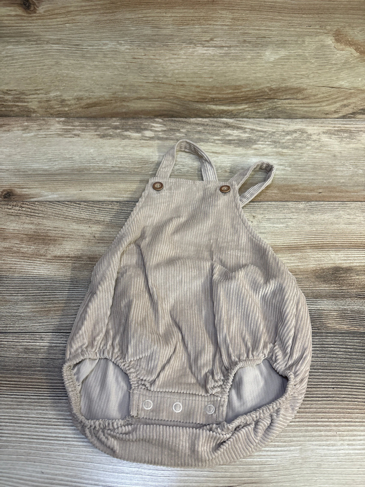 Flat lay of beige corduroy bubble shortalls size 12 months showing wooden strap buttons and elasticized leg openings on wood background