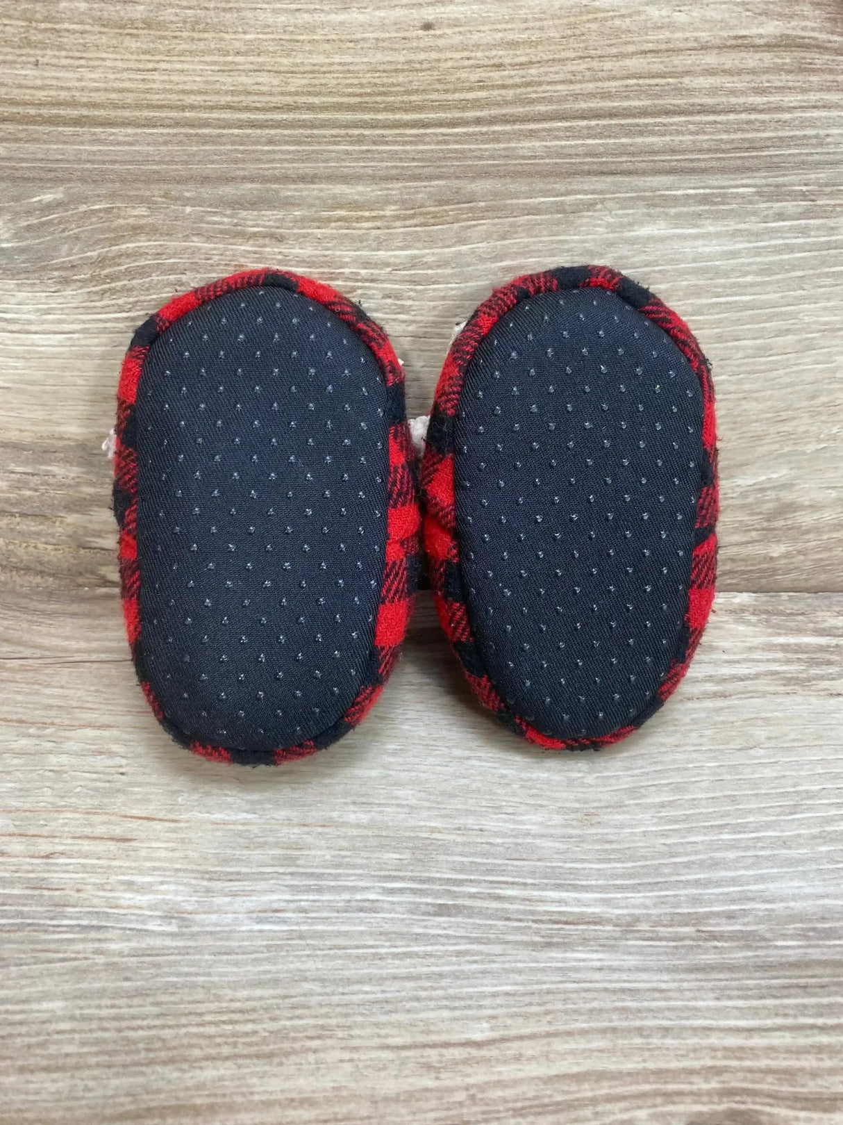 Top view of Buffalo Baby Bear slippers showing black dotted non-slip soles and red plaid trim on wood background