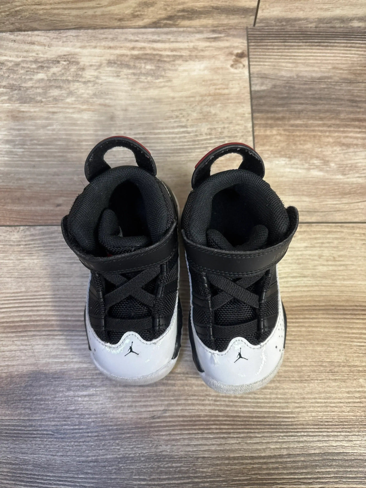 Top view of toddler Air Jordan 6 Rings sneakers with black uppers, white patent toes, Jumpman on toe, and red heel tabs on wood floor