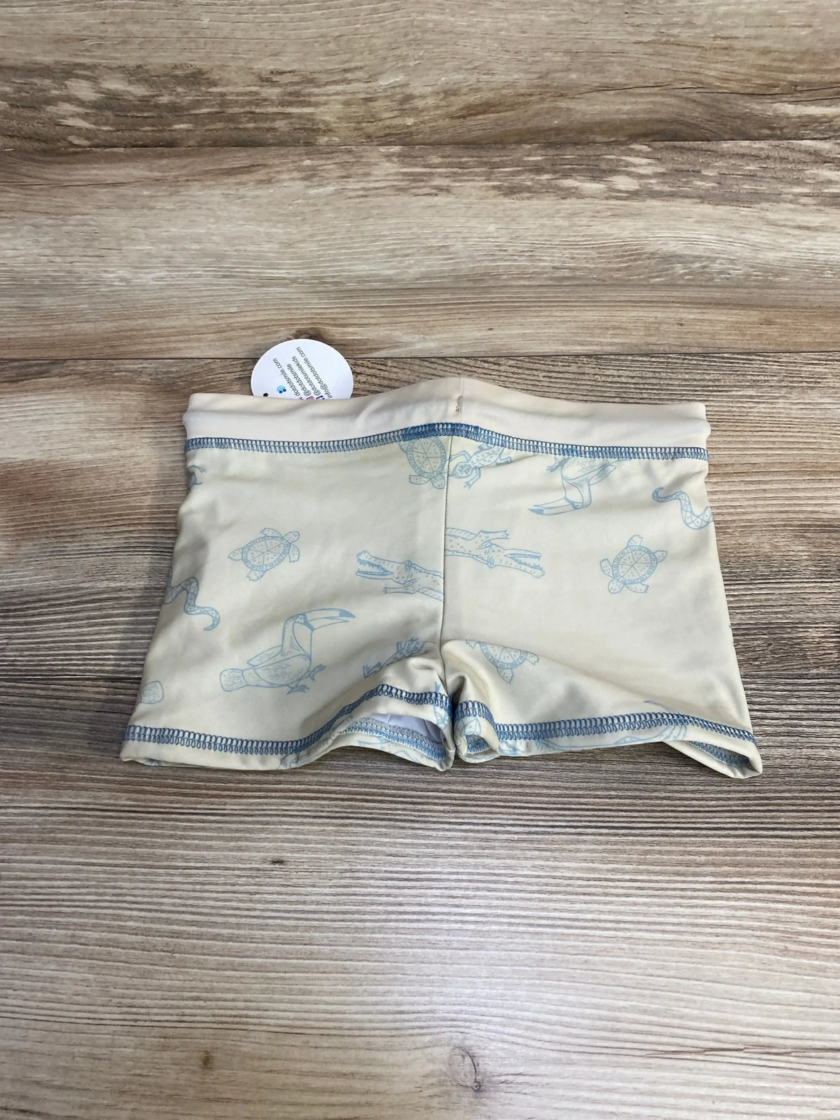 Flat lay of beige toddler swim shorts showing light blue animal prints of turtle, toucan, and snake, blue topstitching on wood background