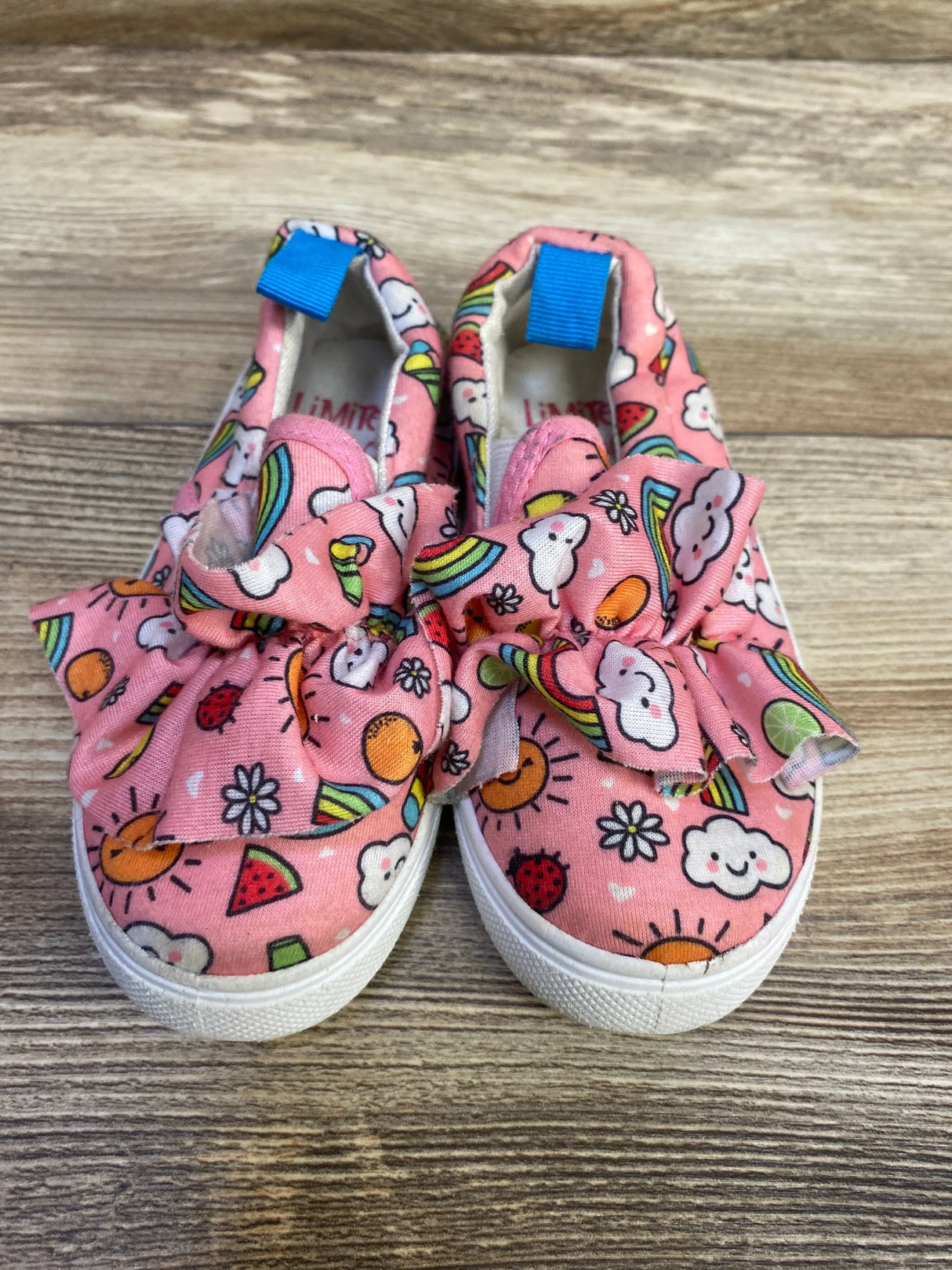 Top-front view of Limited Too pink slip-on sneakers size 8C showing fabric bows, sun and rainbow print, white rubber soles on wood
