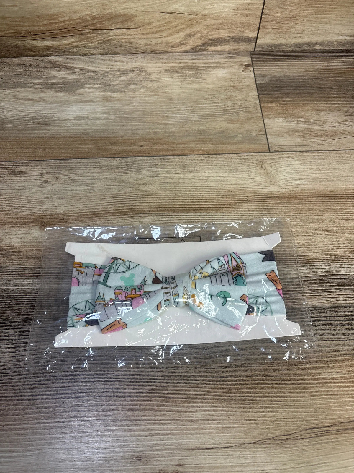 Front view of Disney Park Theme Bow Headwrap showing attached bow and pale blue park-themed print in clear packaging