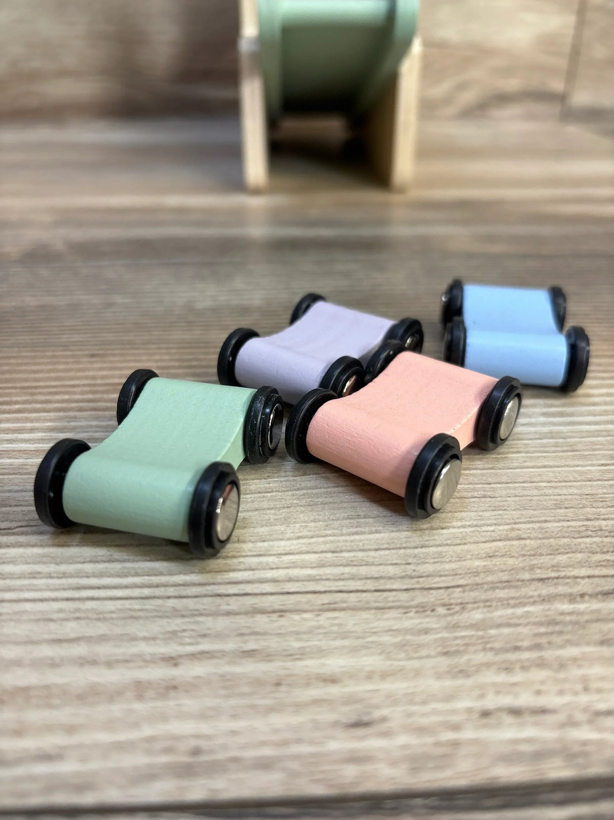 Close-up of Moover Dinosaur wooden race ramp with four pastel cars showcasing black wheels and curved wooden bodies with wooden ramp blurred in background