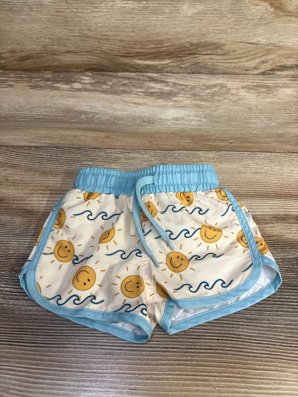 Front view of Caden Lane Sunshine swim trunks with cream background, smiling sun and blue wave print, blue trim on wood floor