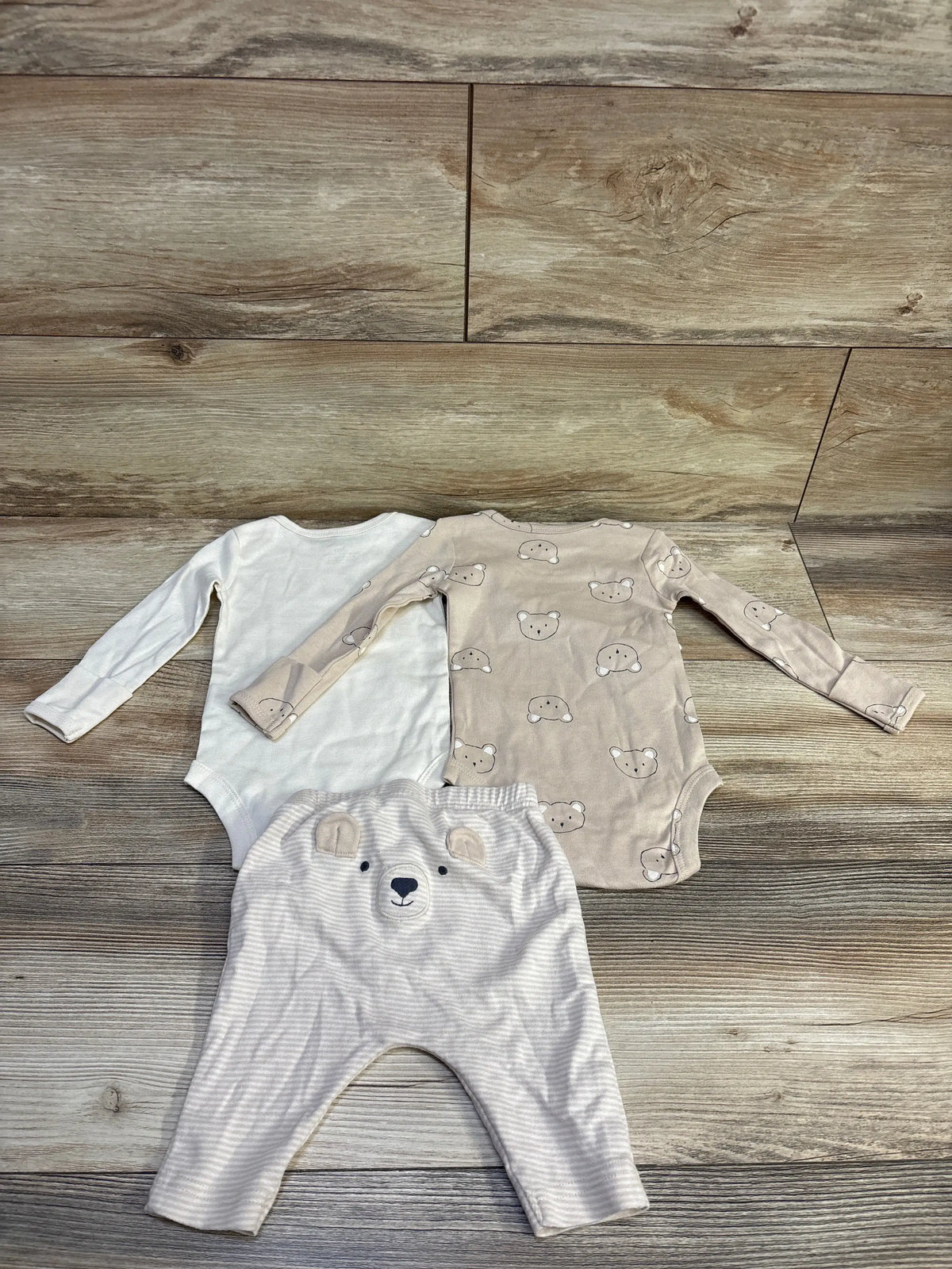 Flat lay of 3-piece long-sleeve bodysuit set: white bodysuit, tan bear-print bodysuit, beige pants with bear face on wood