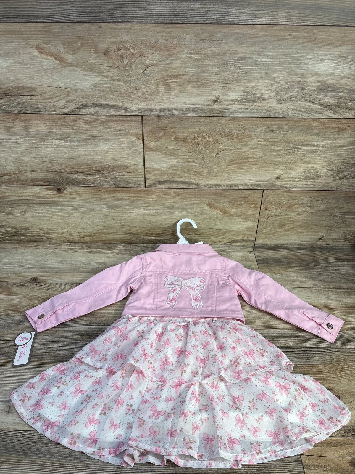 Back view of Little Lass pink two-piece jacket and dress set showcasing bow details and layered skirt on a wood background