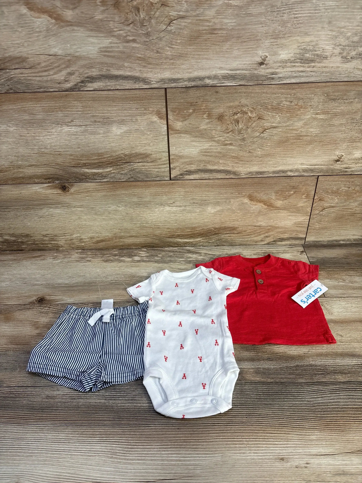 Flat lay of newborn 3-piece set: red Henley top, white onesie with red cherries, navy striped shorts on wood background