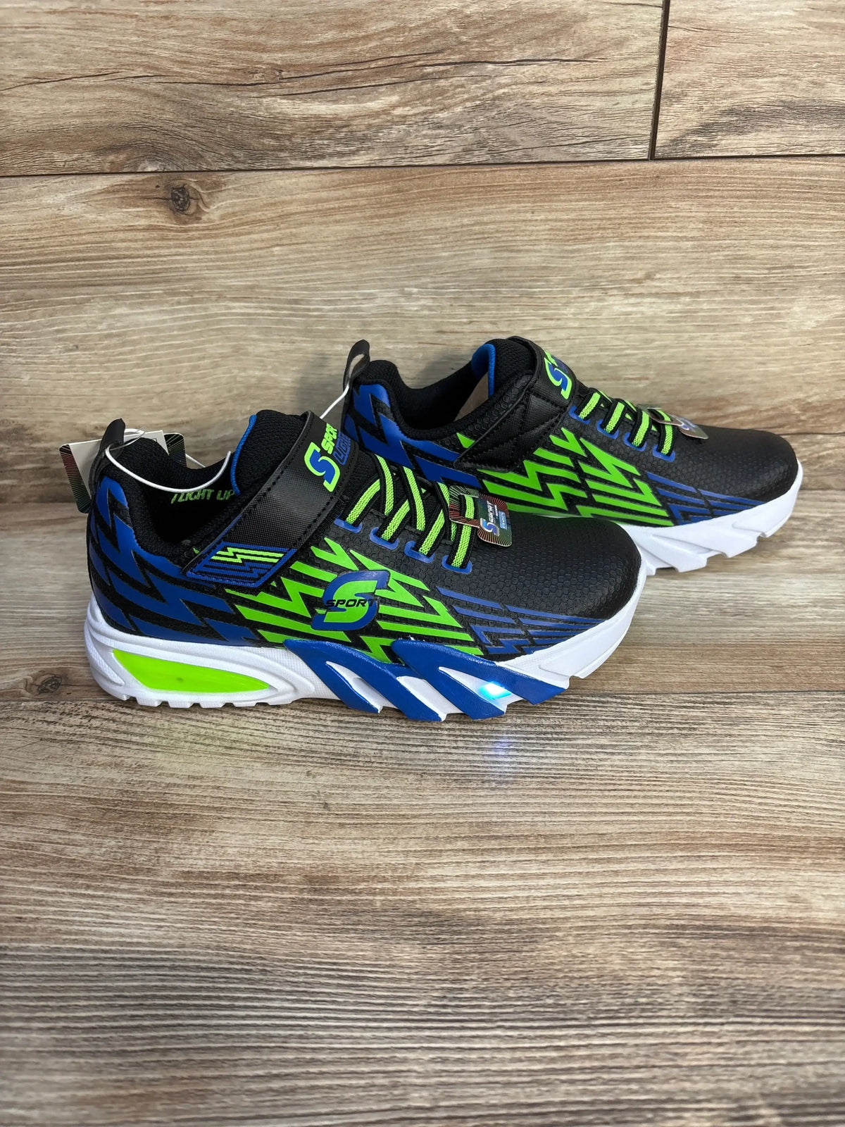 Side view of Skechers Lights Ottis sneakers size 3Y black with blue-green lightning graphics and light-up sole on wood floor