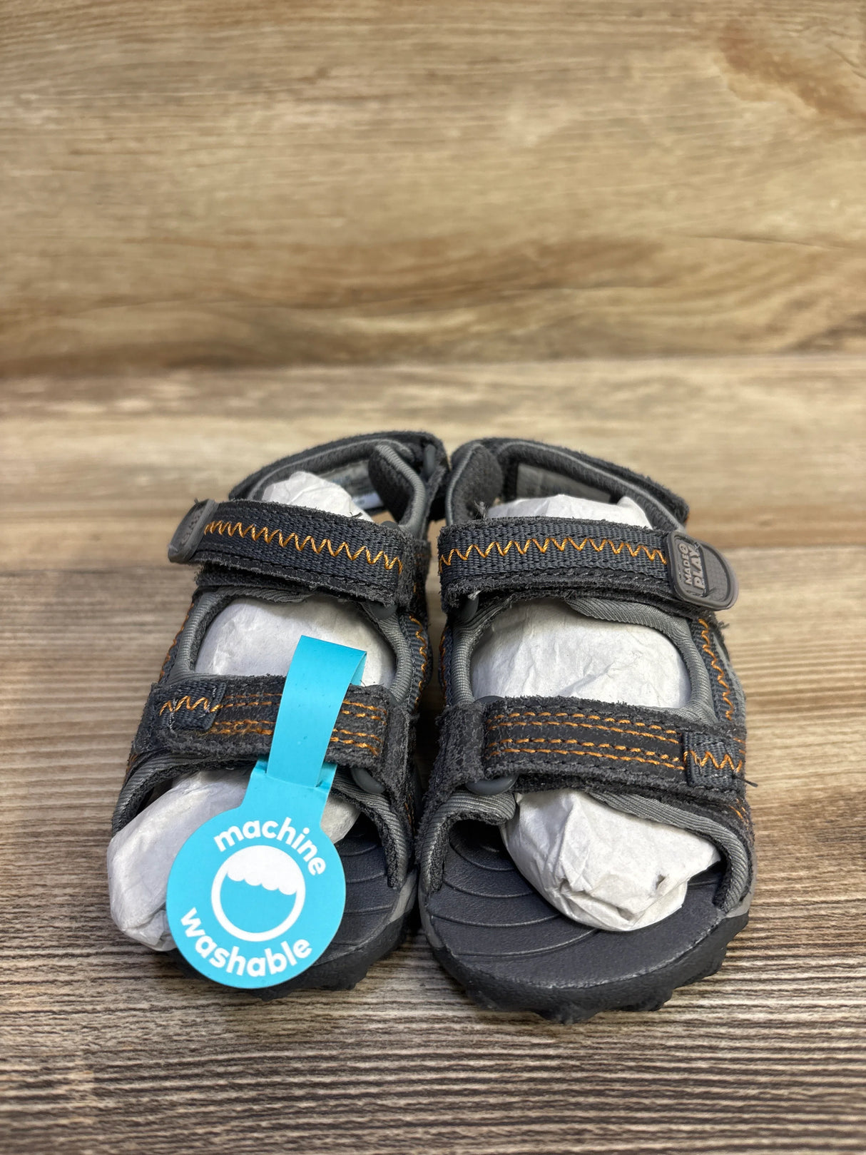 Front view of Stride Rite Made 2 Play Isaac sandals showing grey fabric, orange stitching, Velcro straps and machine-washable tag on wood