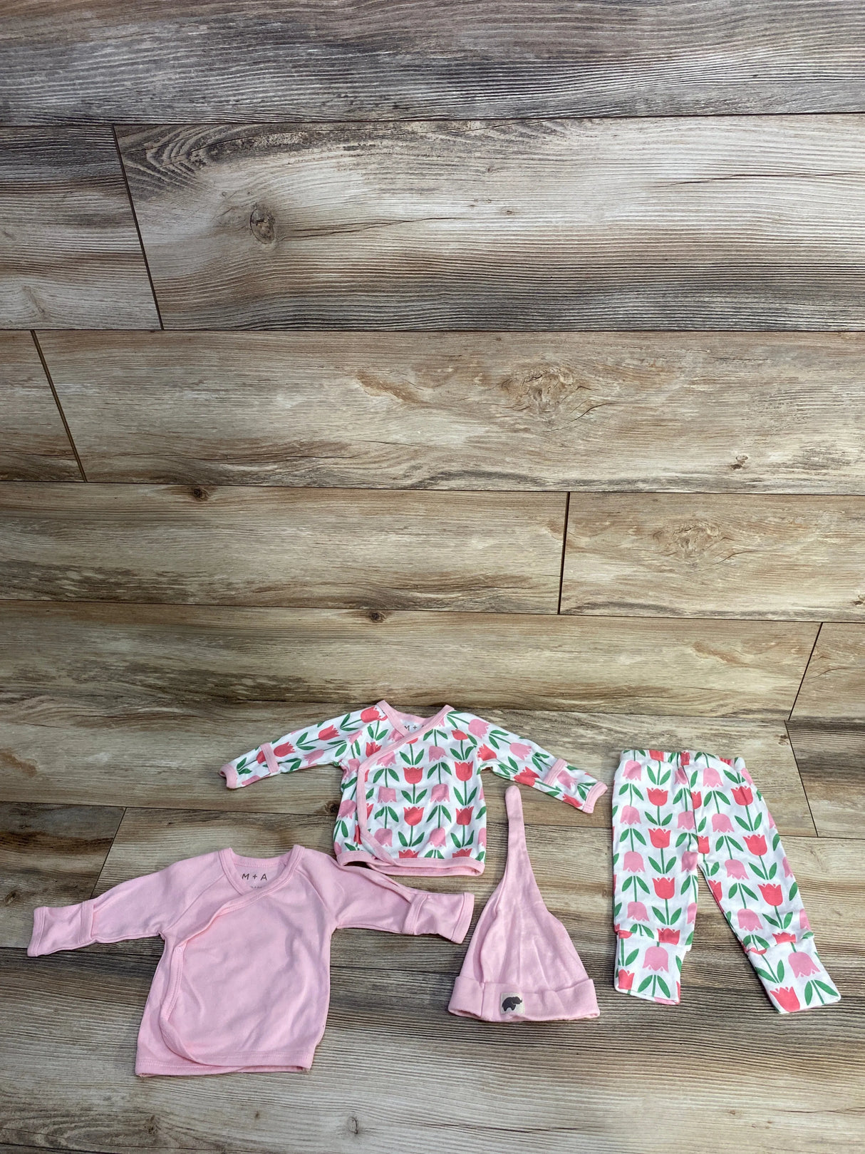 Pink and floral baby clothes set including tops, pants, and a knot hat on wooden floor
