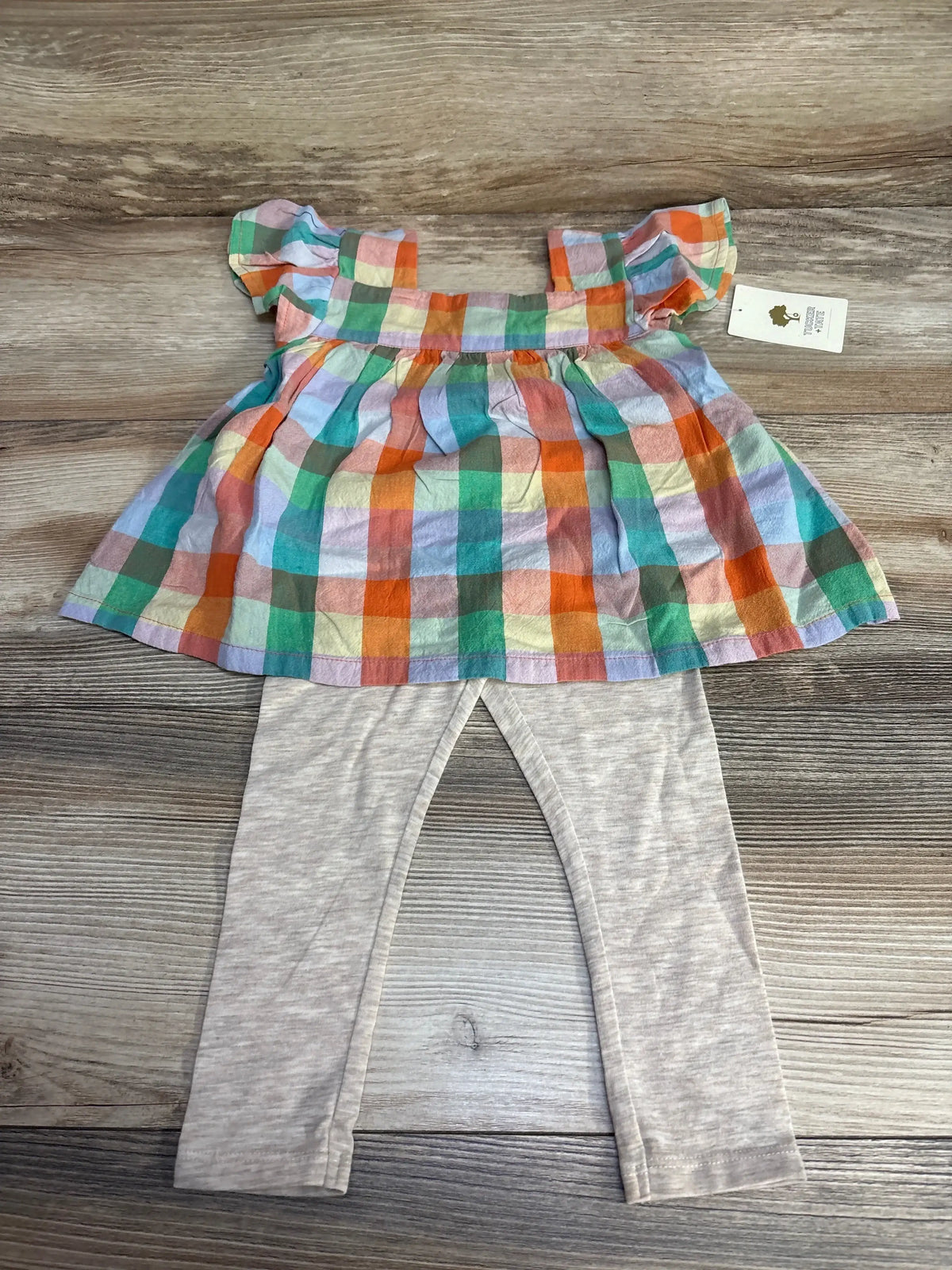 Flat lay of 2-piece toddler set: multicolor checked peplum top with flutter sleeves and light gray leggings on wood background