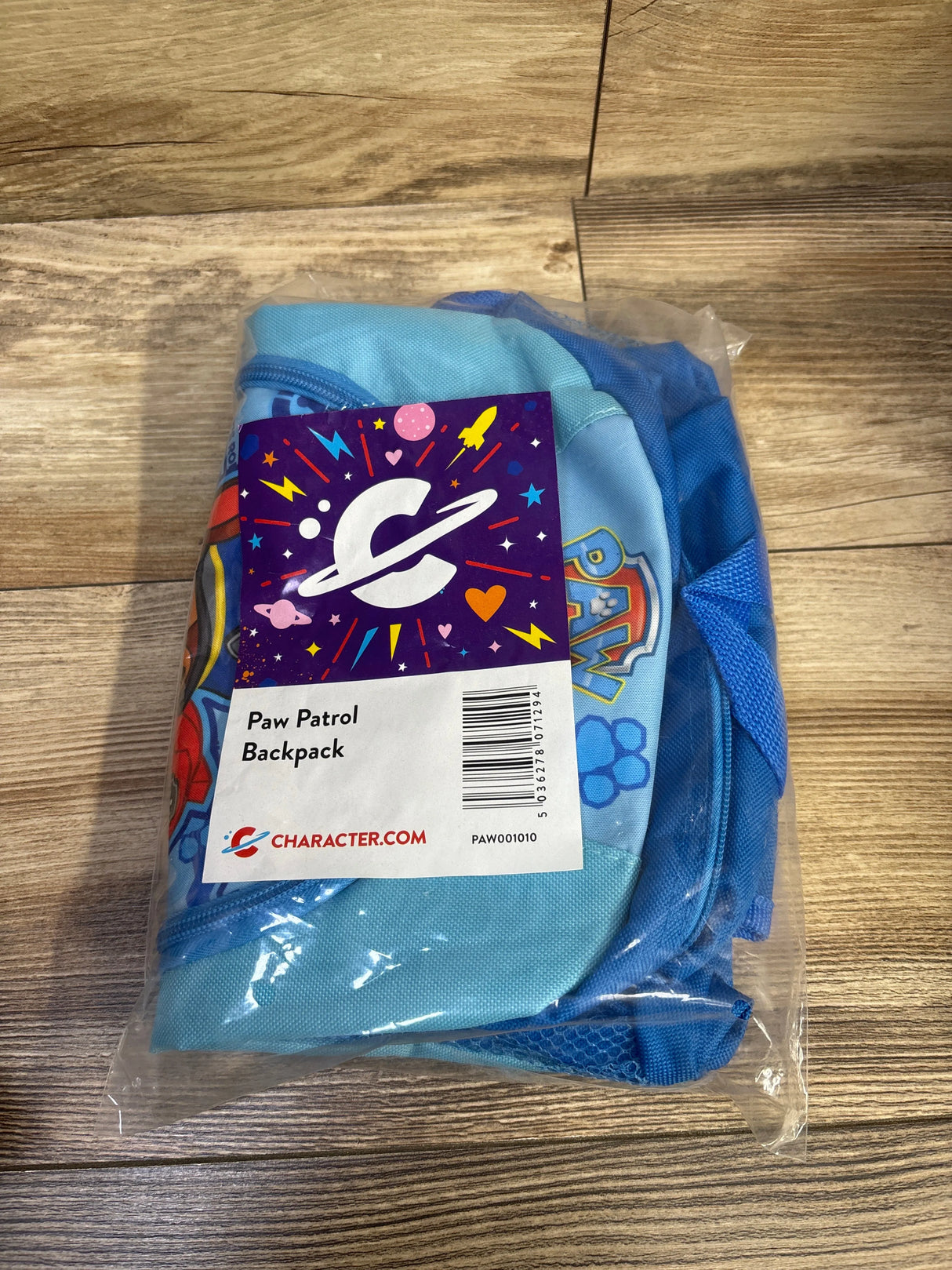 Front view of Paw Patrol kids backpack in clear packaging, blue color with front logo patch and mesh side pocket on wood tabletop