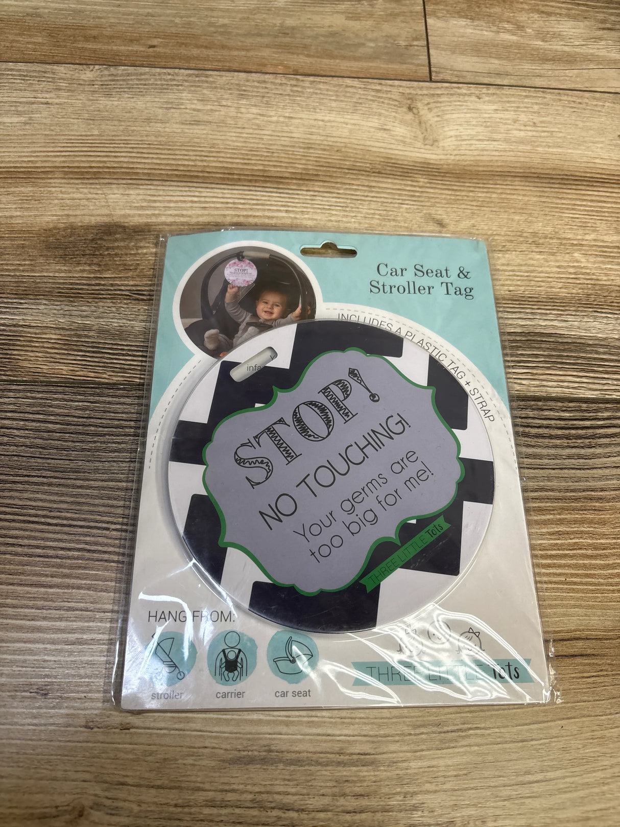 Top-down view of a round car seat and stroller tag showcasing the message STOP! NO TOUCHING! Your germs are too big for me with a wooden floor background
