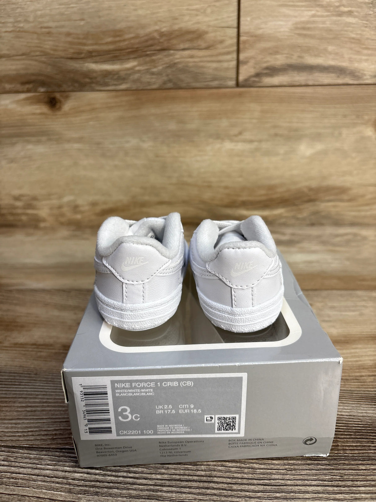 Pair of white Nike Force 1 Crib baby shoes on gray Nike shoebox with wooden background