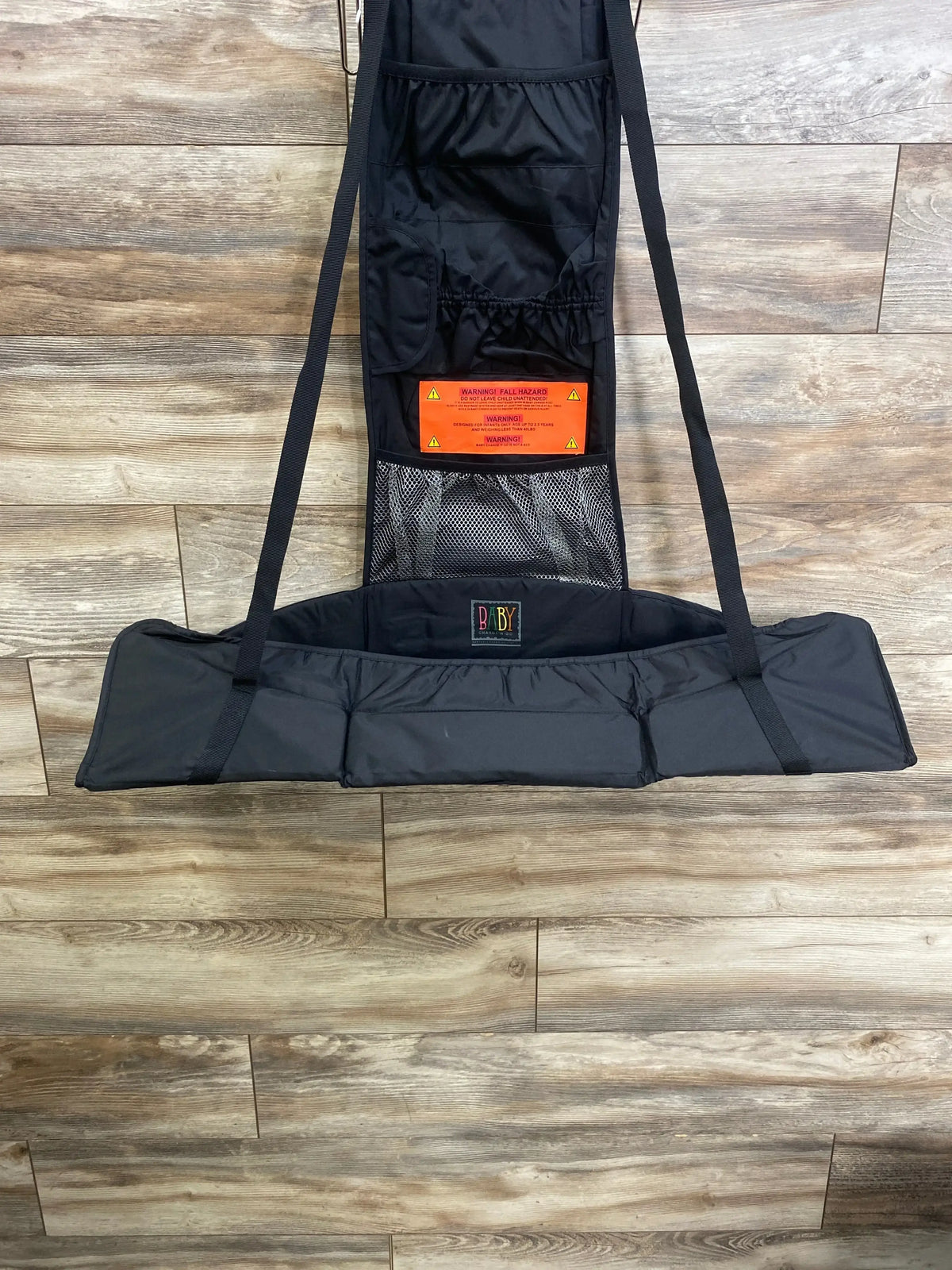 Front view of Baby Change-N-Go portable hanging changing station with padded surface, mesh pocket, and straps on wood-look floor