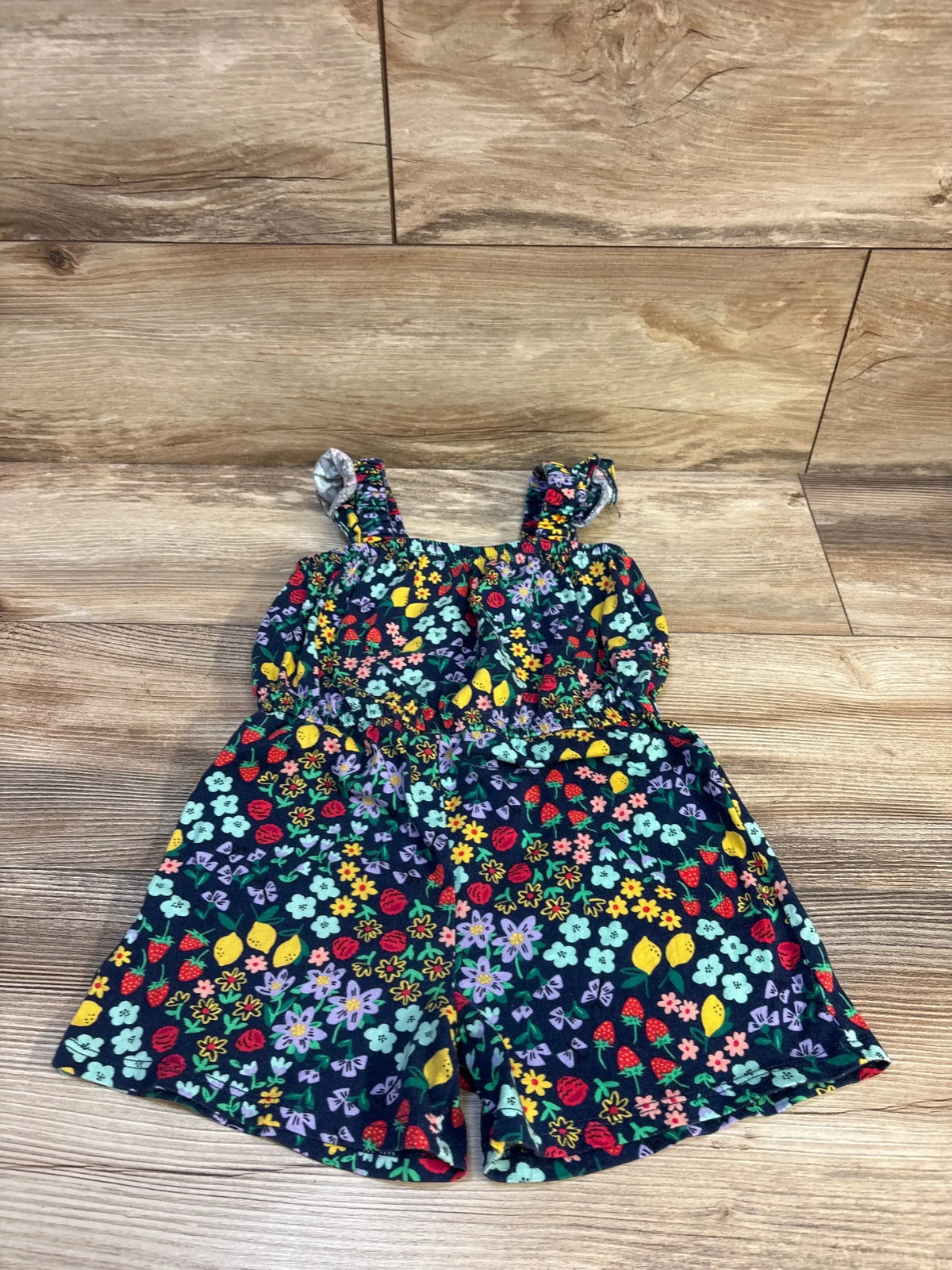 Flat lay of navy sleeveless romper showing fruit and floral print, ruffled straps and elastic waist on wood floor
