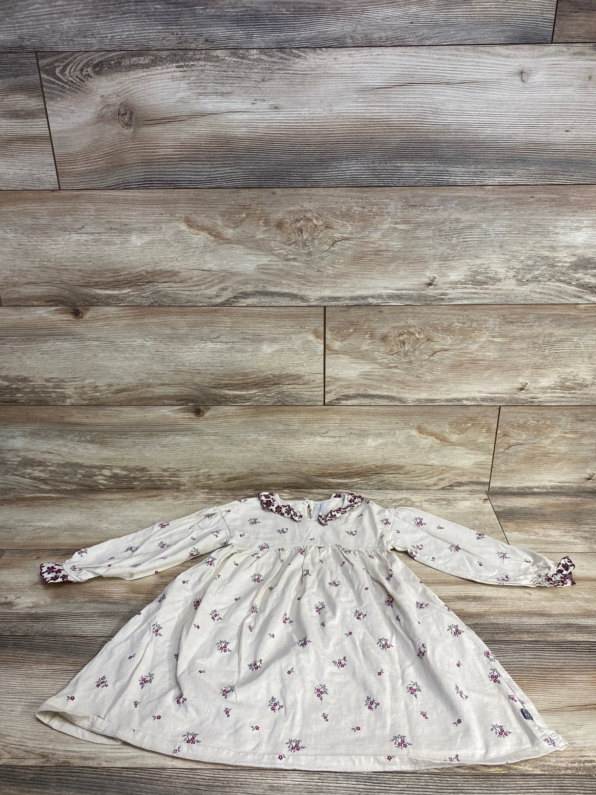 White long-sleeve baby dress with purple floral print and ruffled collar, laid flat on wood floor