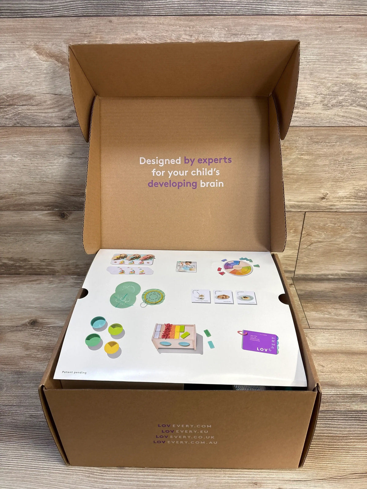 Top-down view of Lovevery The Analyst Play Kit open box showcasing illustrated activity cards, wooden sorting toys, and packaging on a wood floor