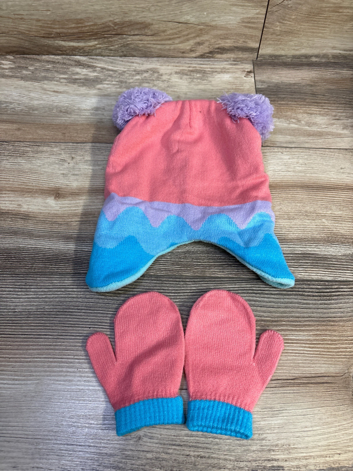 Flat lay of Abby Cadabby hat and mittens set: pink hat with purple pom-pom ears and blue wave trim, matching mittens on wood