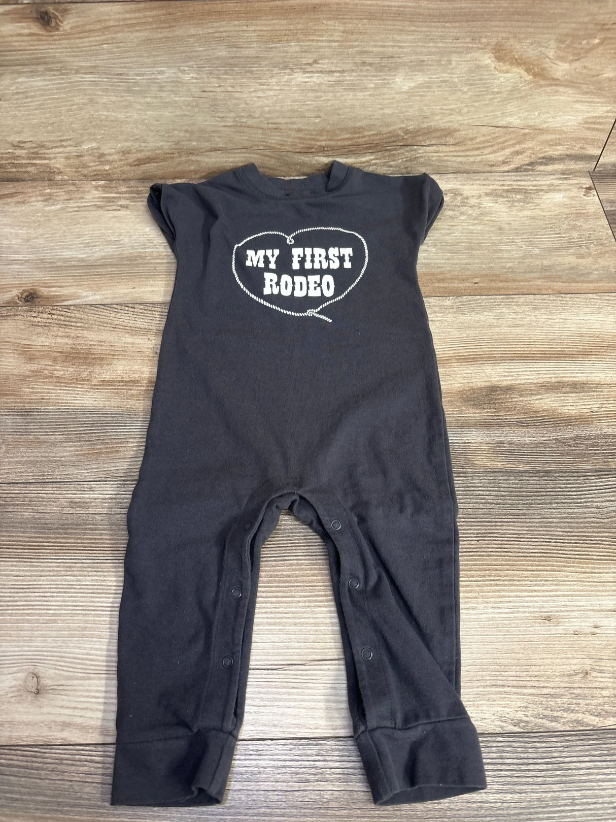 Flat lay of Cat and Jack My First Rodeo Romper in dark gray sz 6-9m showcasing white heart-shaped graphic and snap leg closure on wood plank background