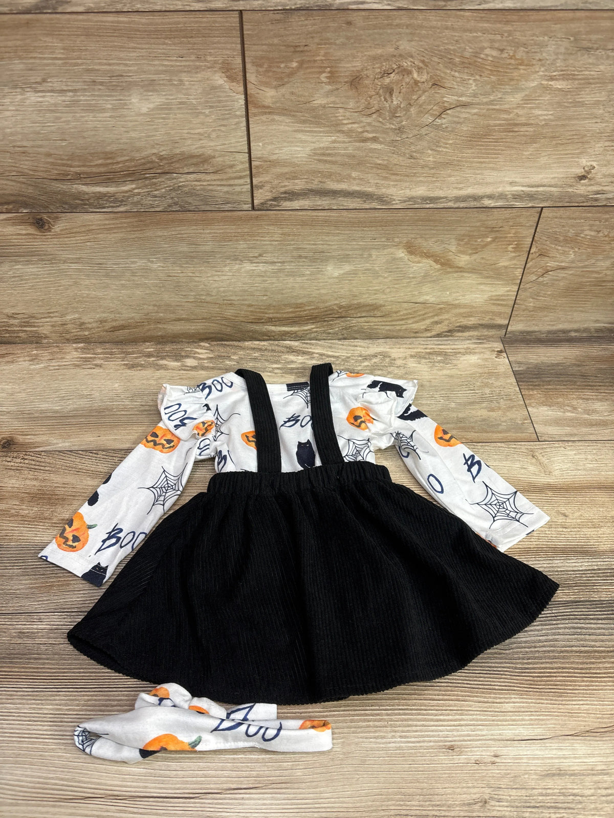 Top-down view of a three-piece Halloween skirtall set with black corduroy skirt, white pumpkin-print top, and matching headband