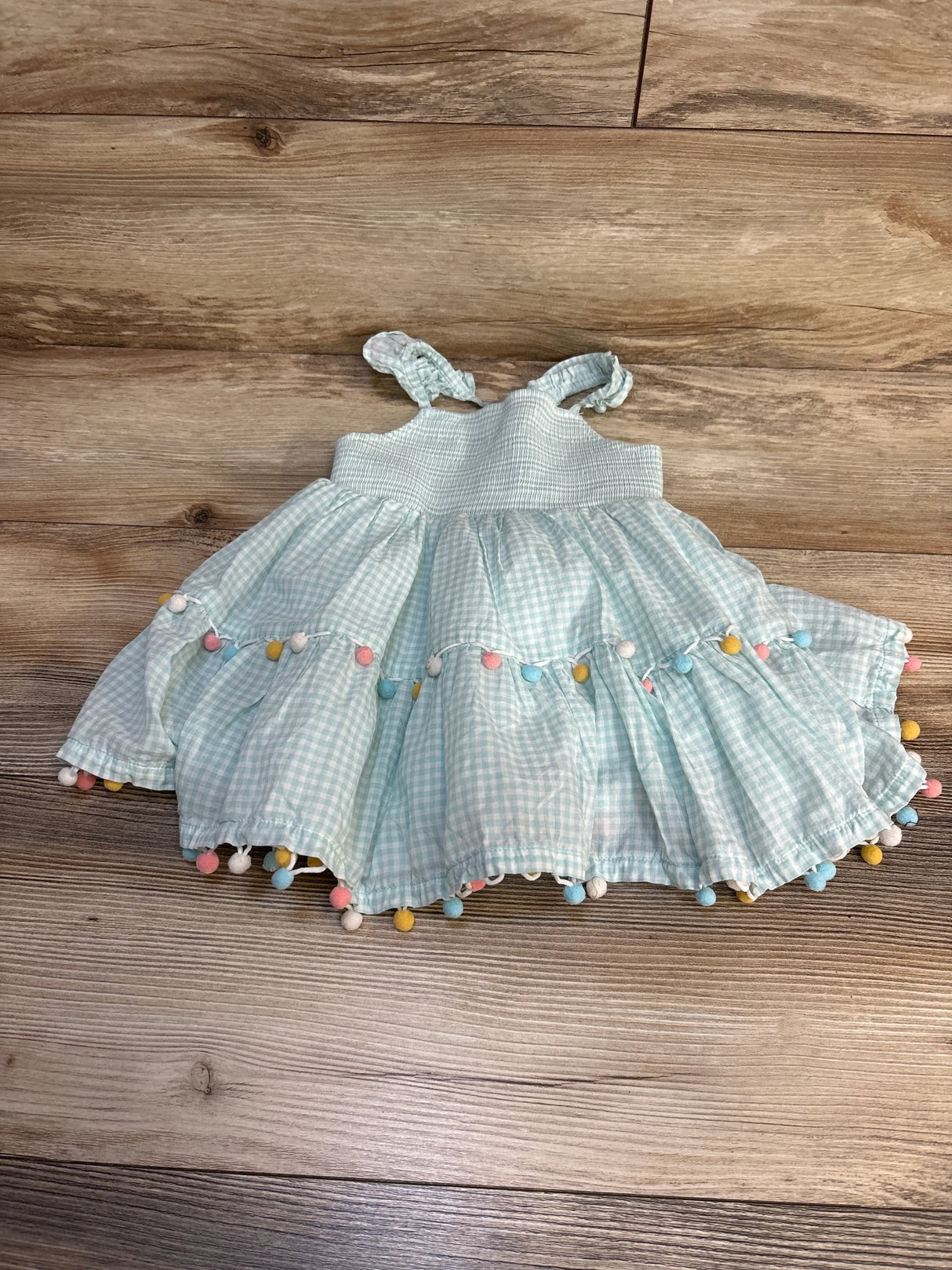 Light blue gingham baby dress with colorful pom-poms on wooden floor